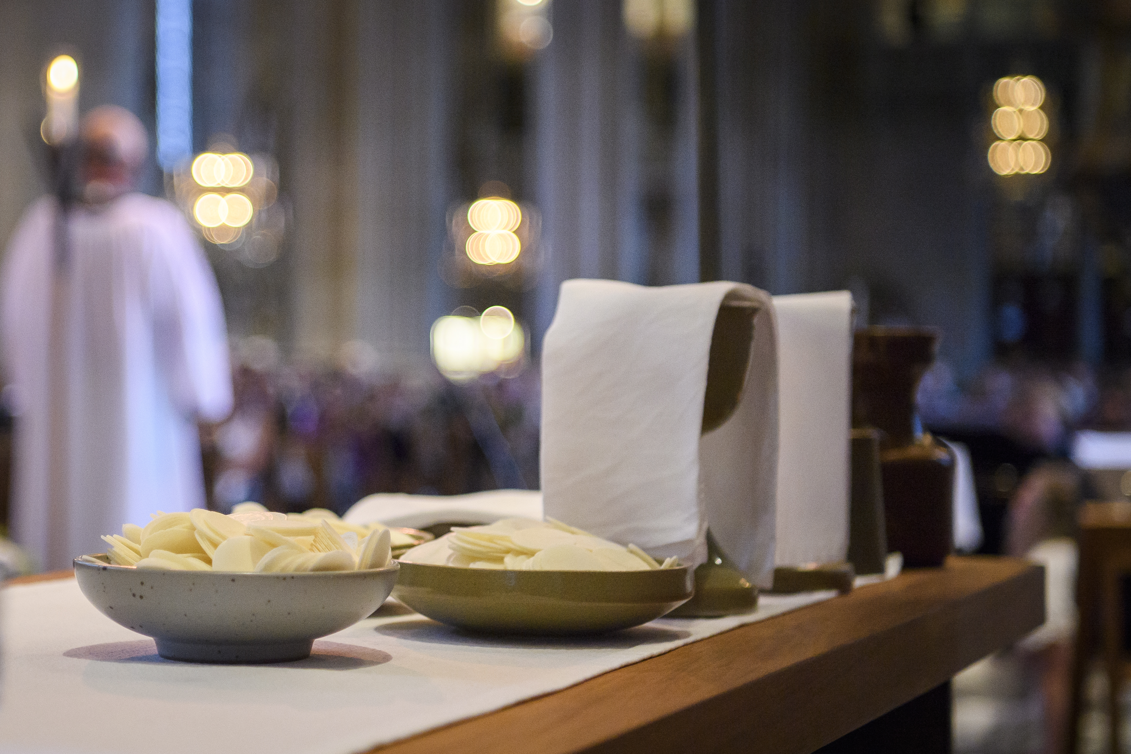 Nattvardsvin och oblat står uppdukat på ett bord. En präst och en många kyrkobesökare skymtas i bakgrunden.