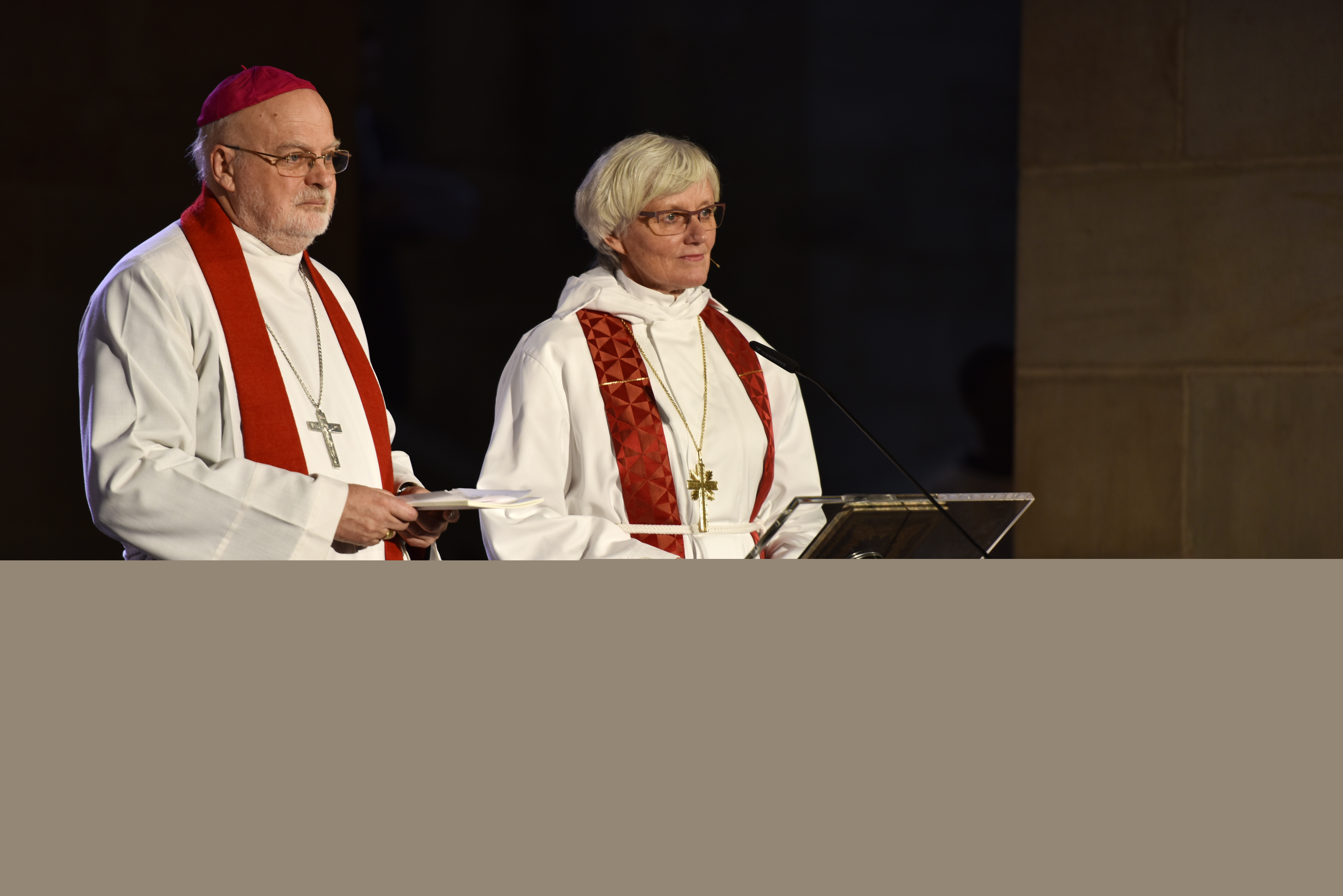 Biskoparna Anders Arborelius och Antje Jackelén står i strålkastarljus i en kyrka och talar i en mikrofon.