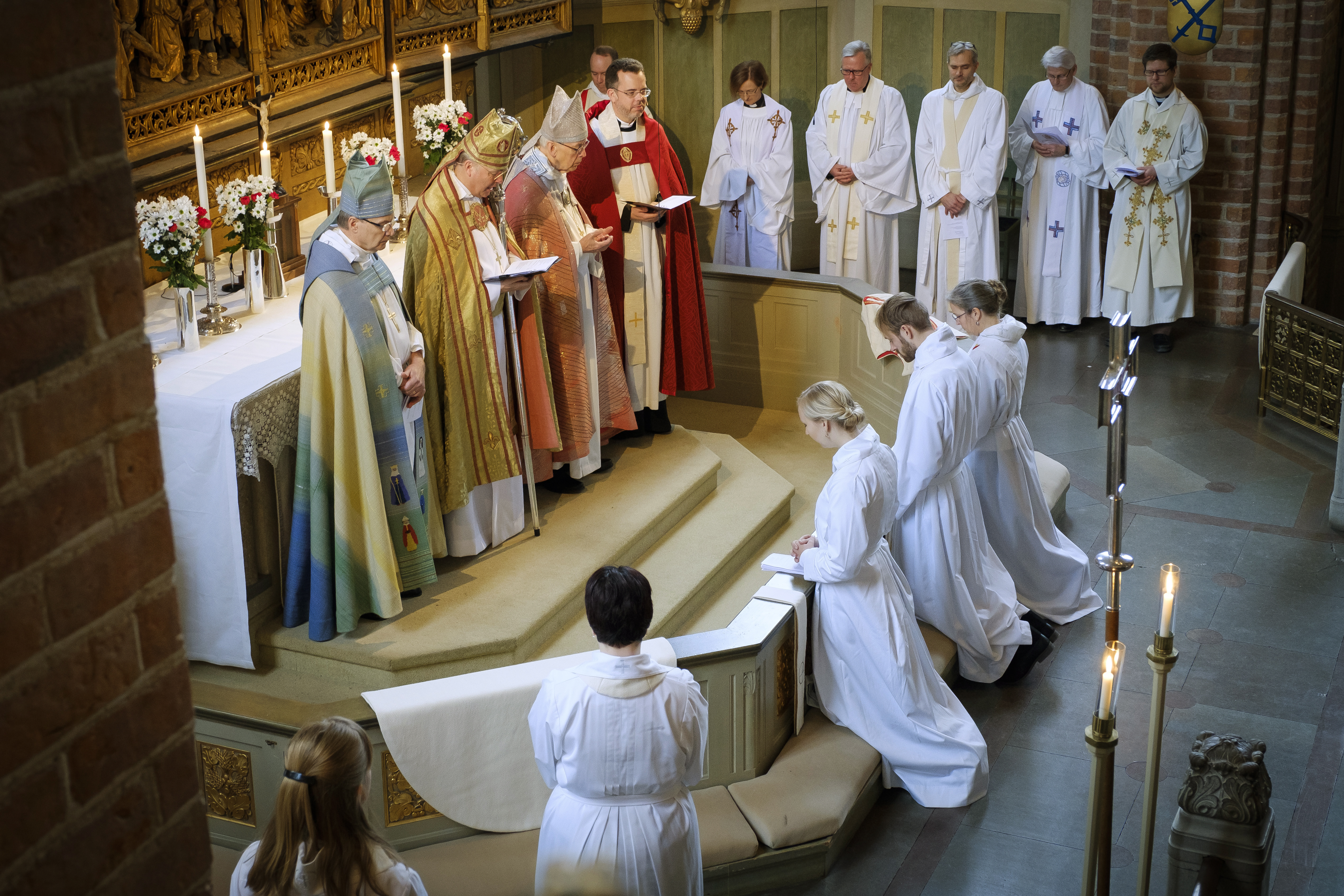 Fyra biskopar står vid ett altare, vända mot tre vitskrudade personer som knäböjer.