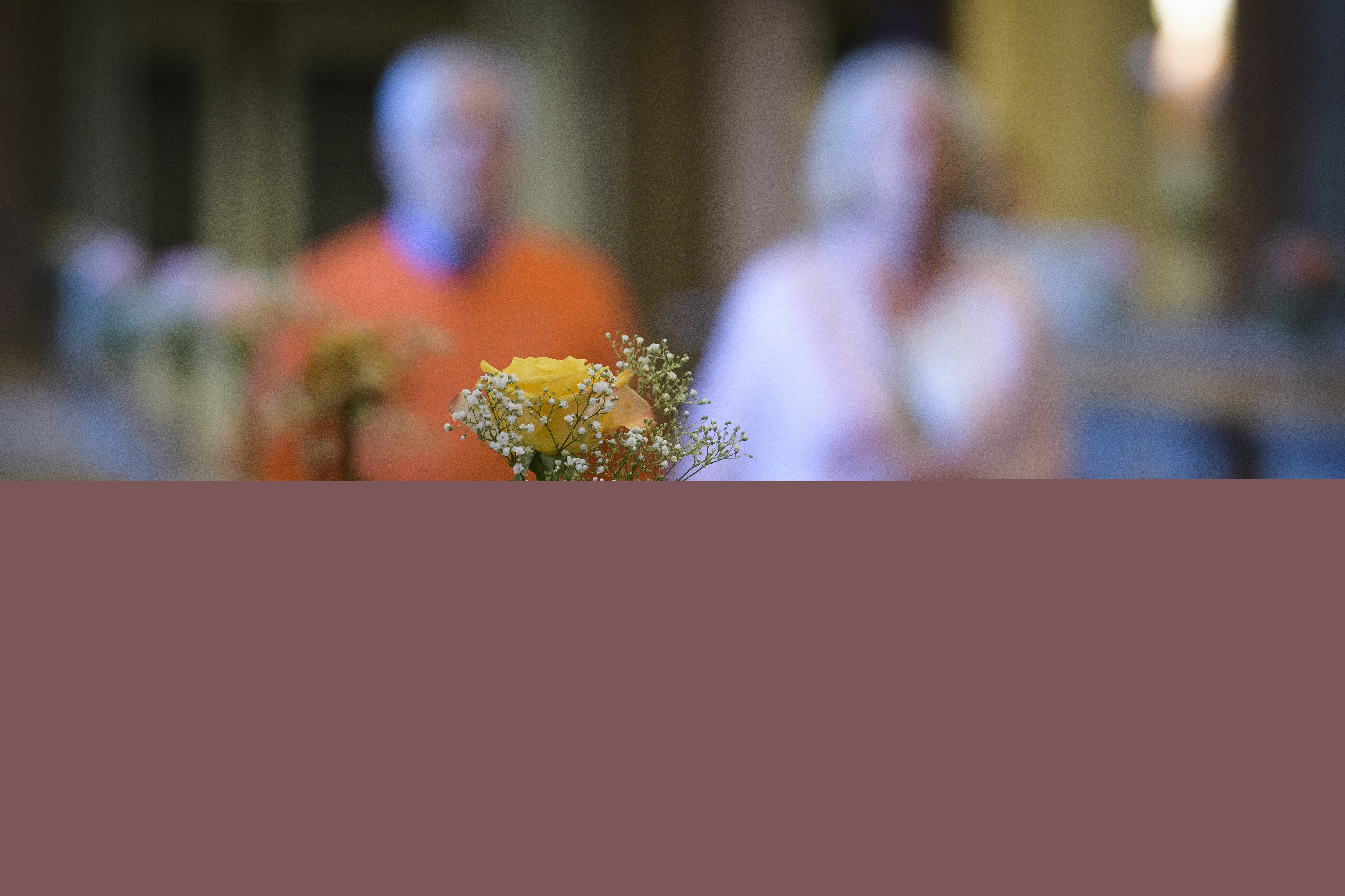 Små vaser med rosor står på bänkraderna i en kyrka. En man och en kvinna syns suddigt gå hand i hand i mittgången.