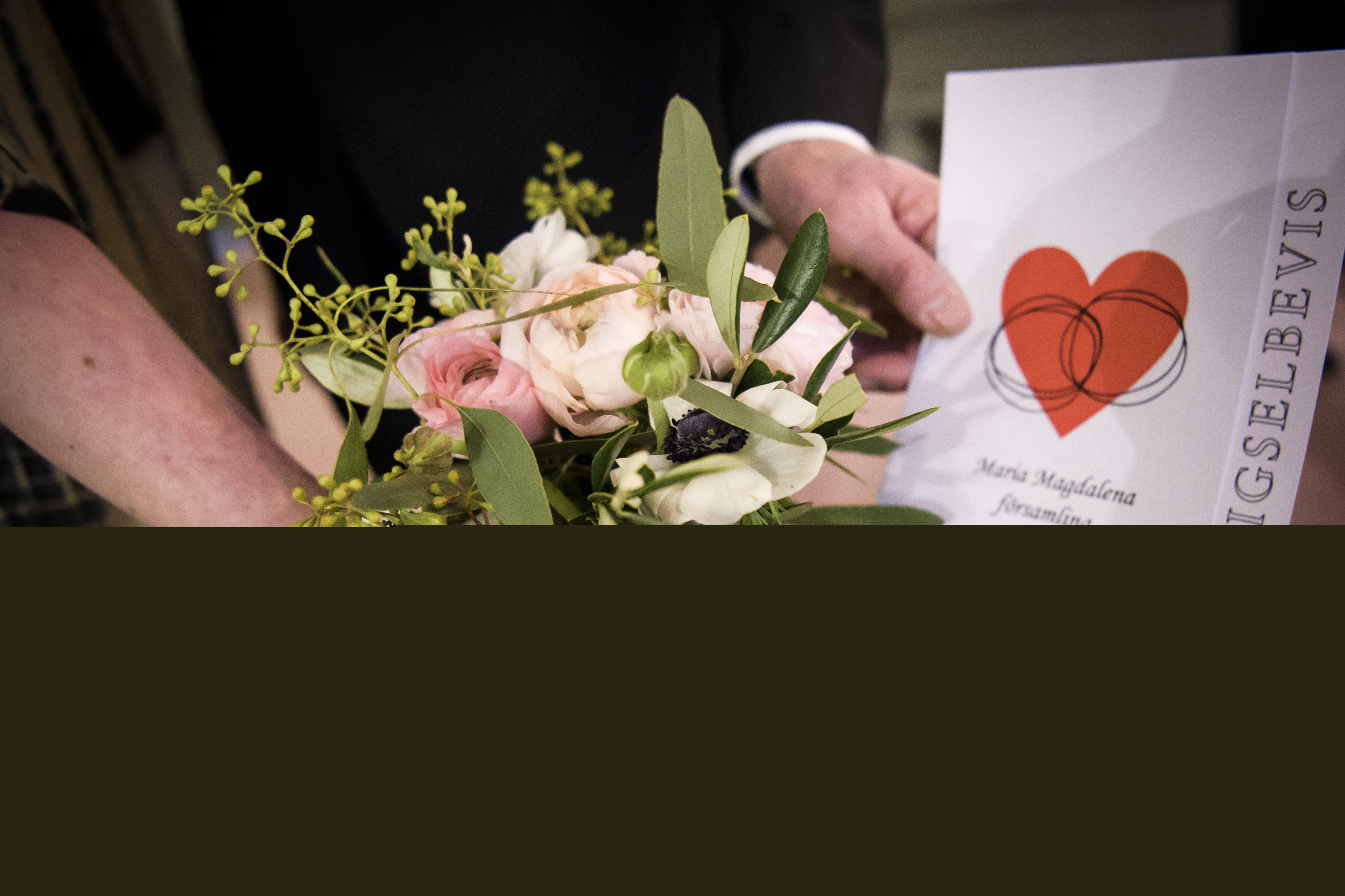 En brud och brudgum håller fram en brudbukett och ett vigselbevis mot kameran.