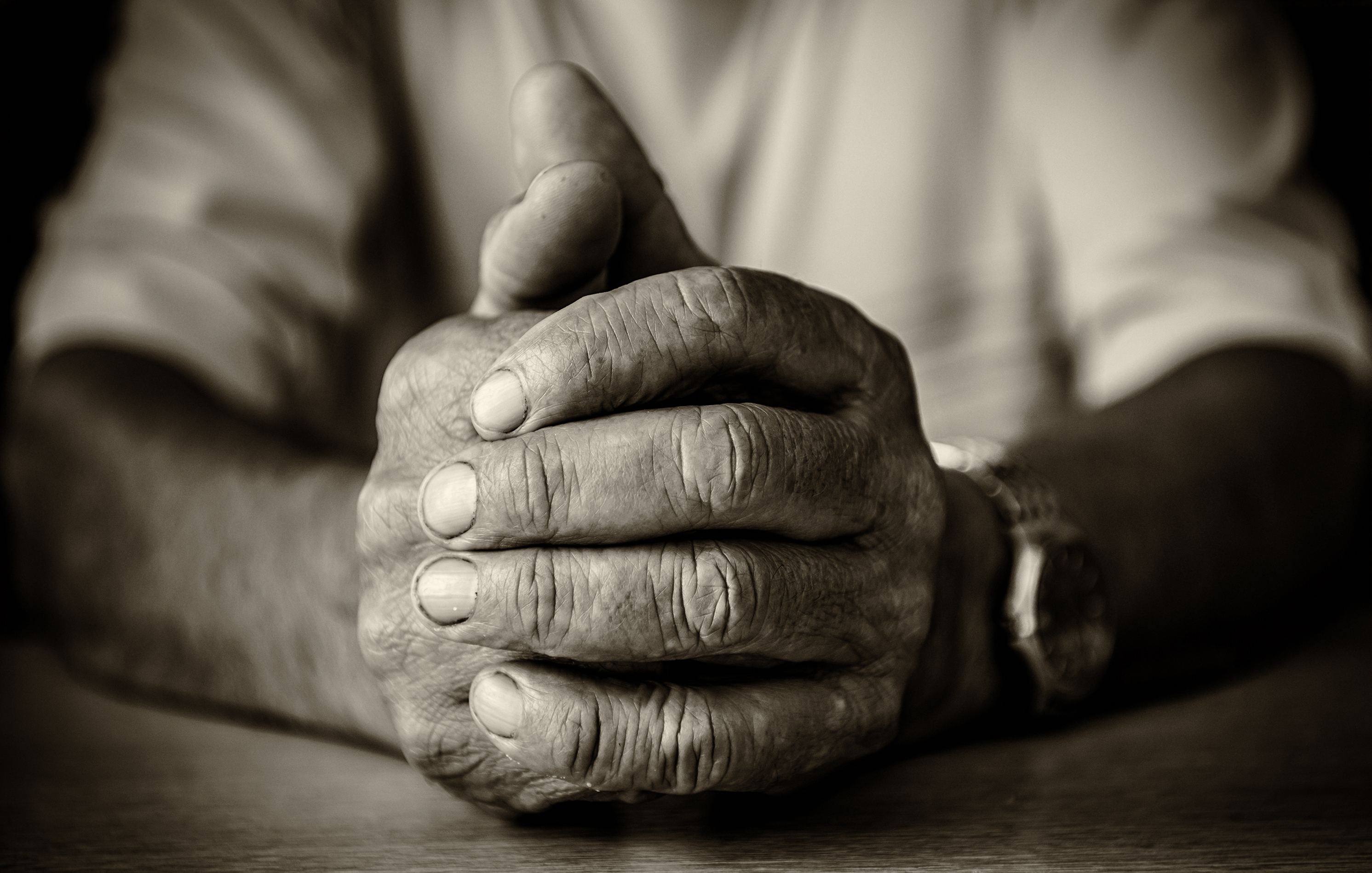 En svartvit bild på en gammal mans händer. Den högra handen håller om den vänstra. Händerna vilar mot ett bord.