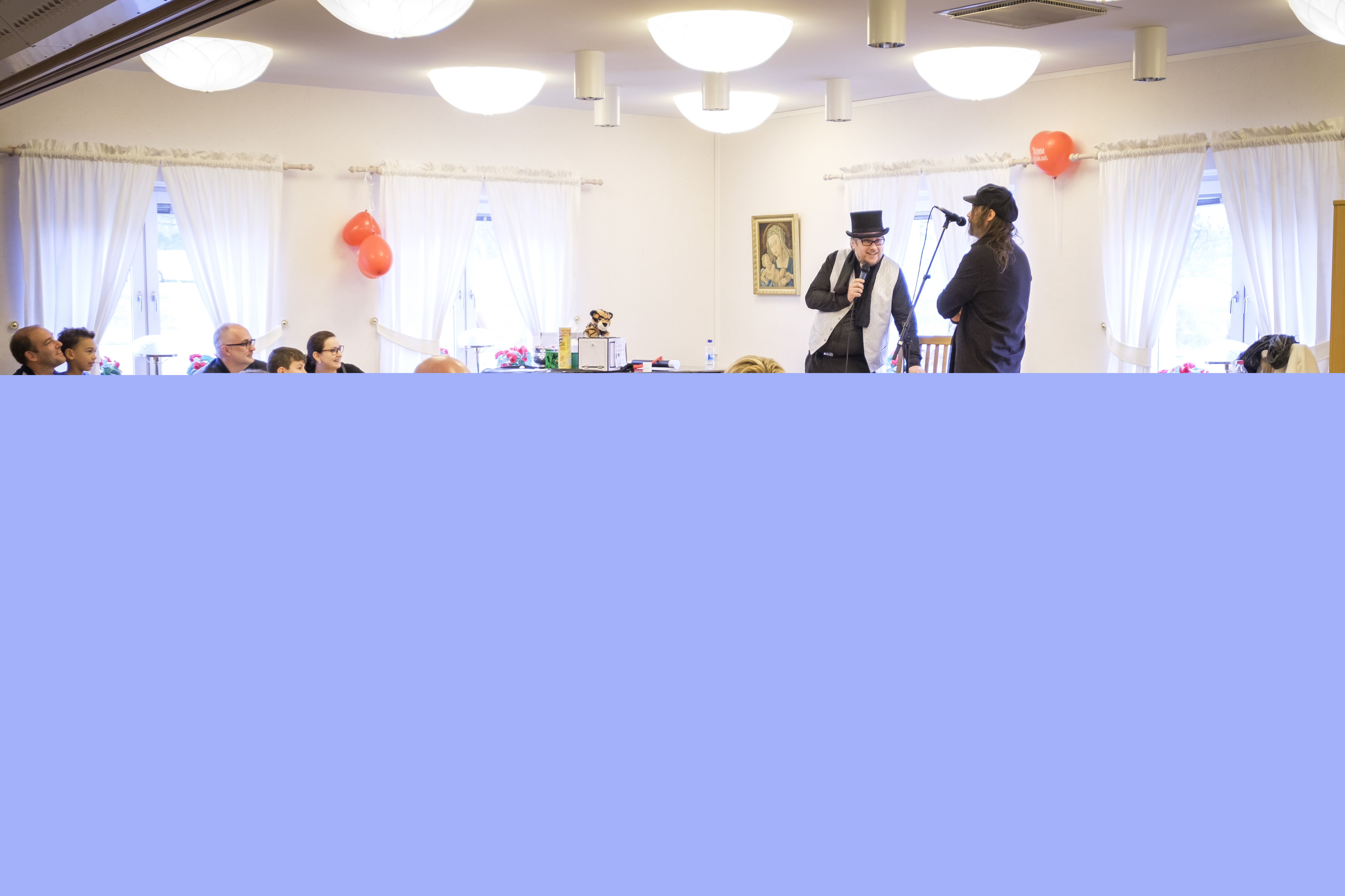 En grupp vuxna och barn sitter i en sal och fikar. I bakgrunden står två musiker på en liten scen.