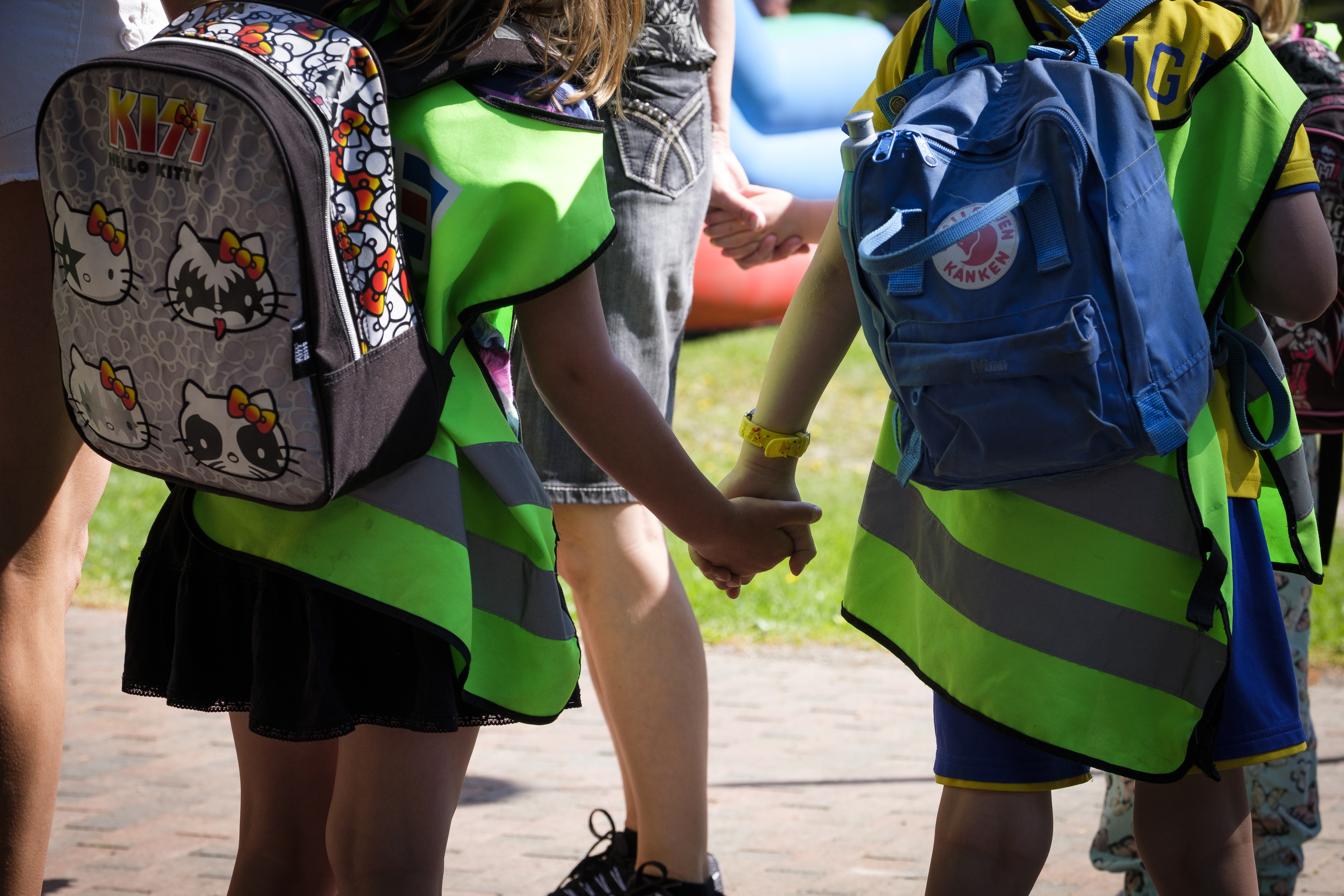 Två småbarn med ryggsäckar och reflexvästar går och håller hand utomhus.