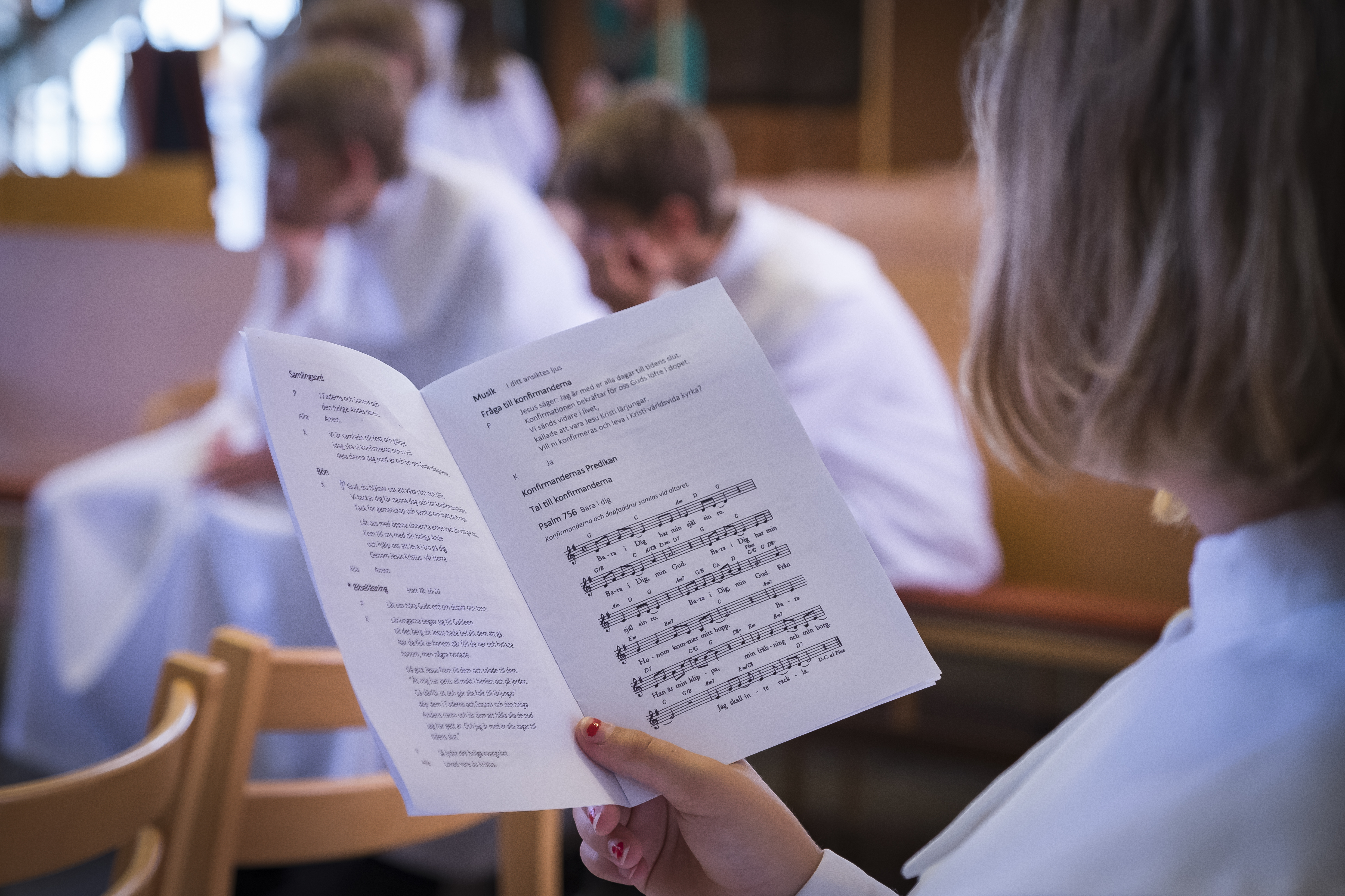 En konfirmand läser i ett sånghäfte. Fler konfirmander sitter i bakgrunden.