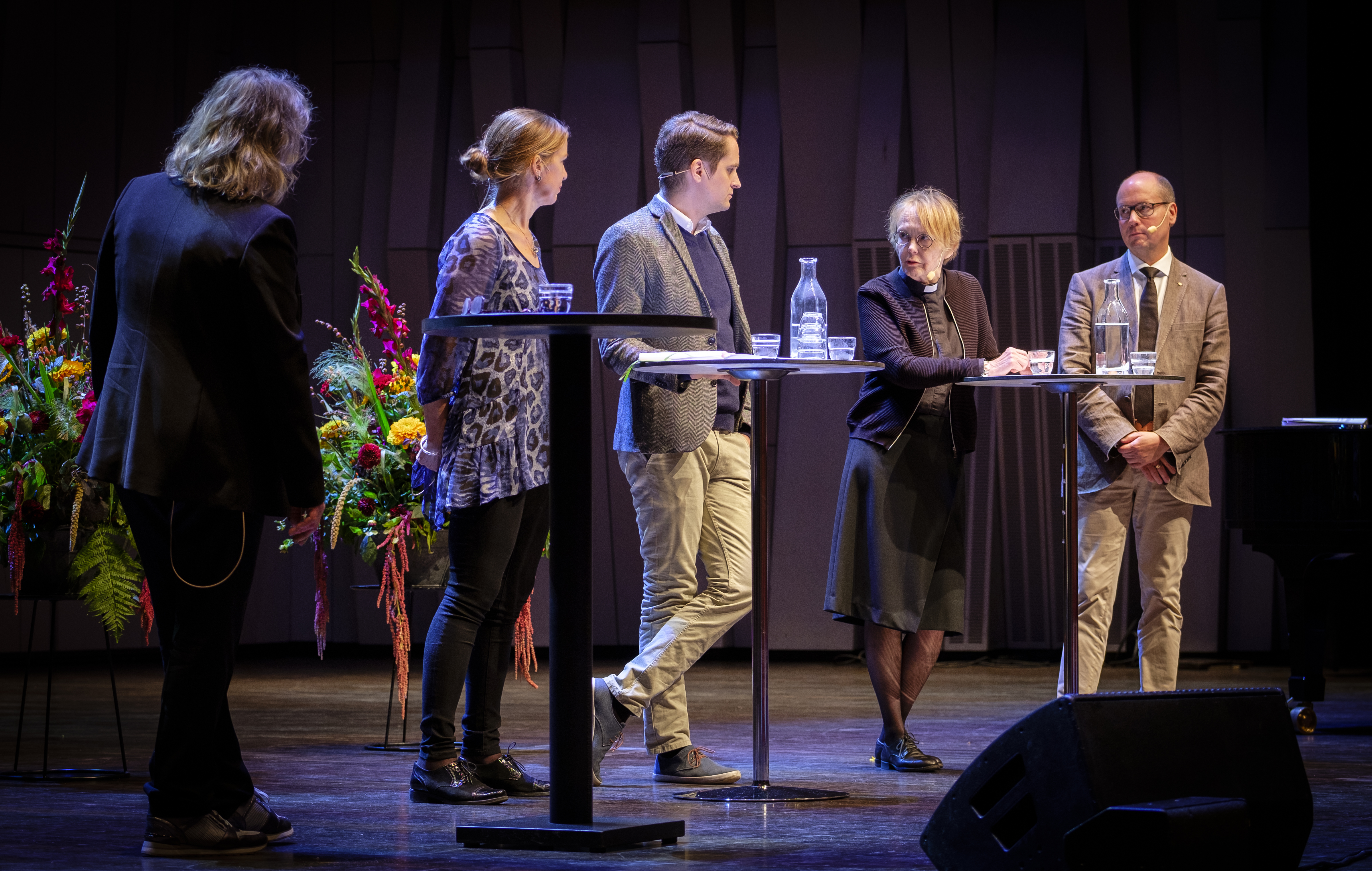 Några personer står runt små bord och pratar på en ljussatt scen.