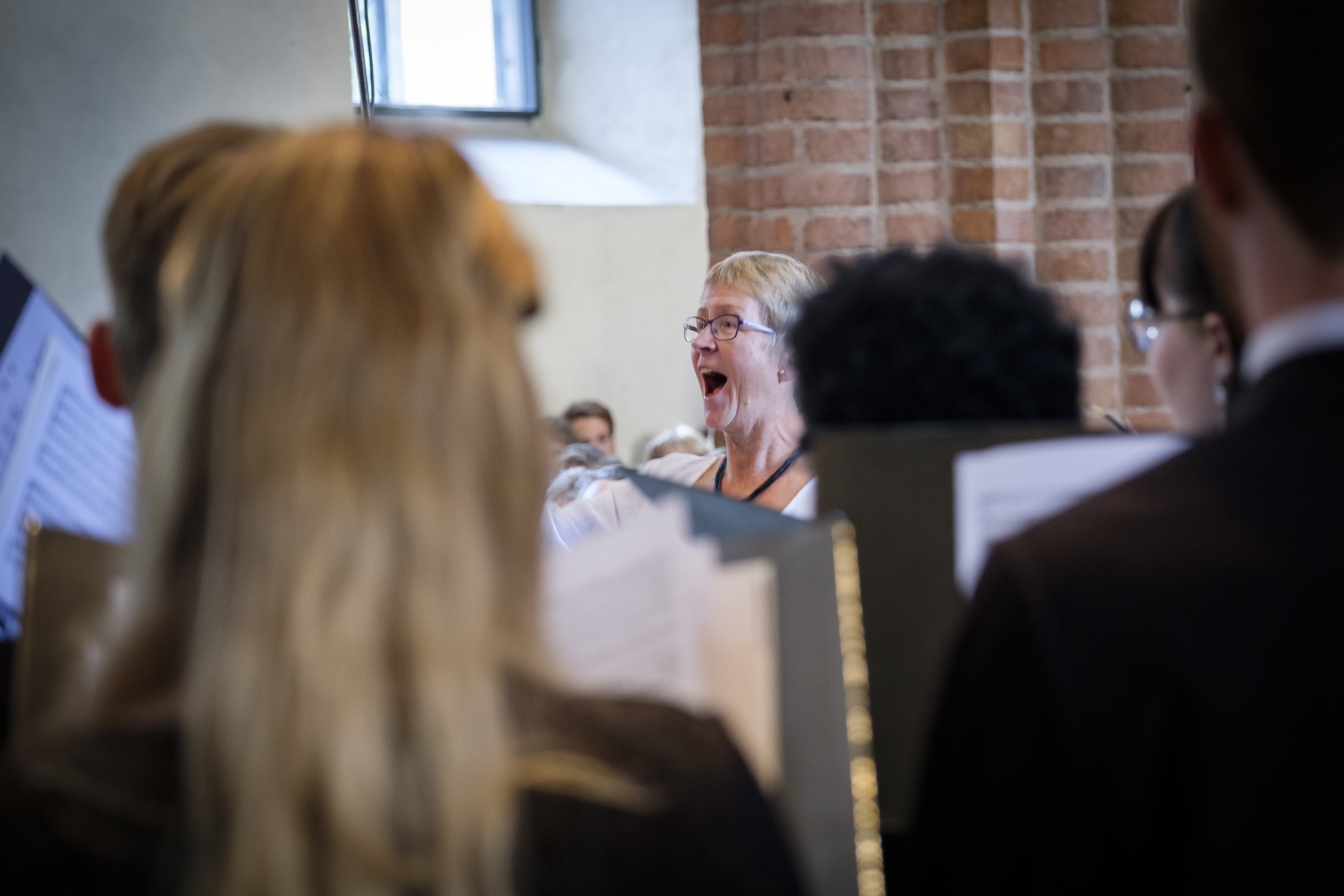 En kvinnlig körledare står i en kyrka och dirigerar. Körmedlemmar i förgrunden.