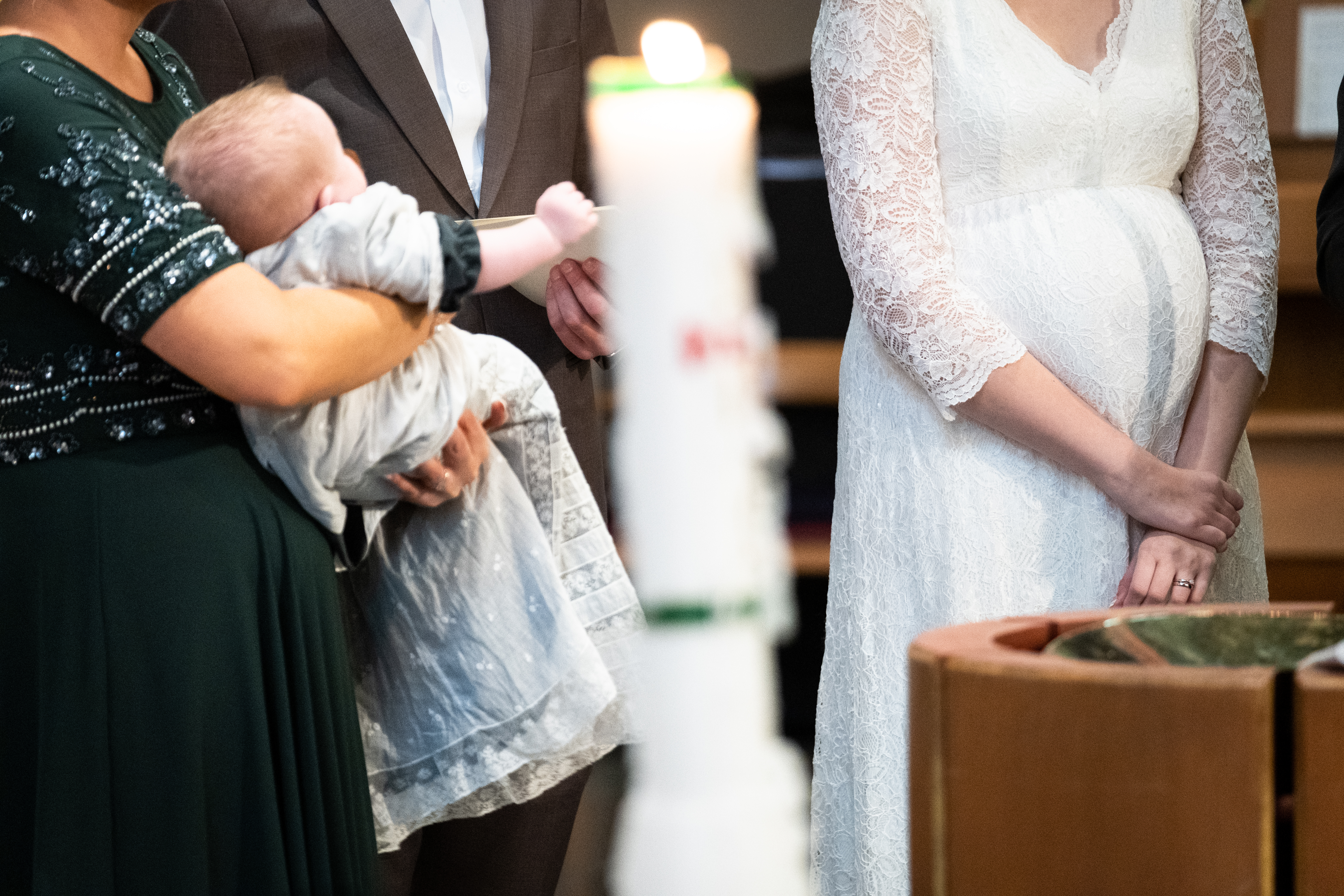 En kvinna i grön klänning bär på en bebis i dopklänning. Bortom dopfunten står ett par i bröllopskläder.