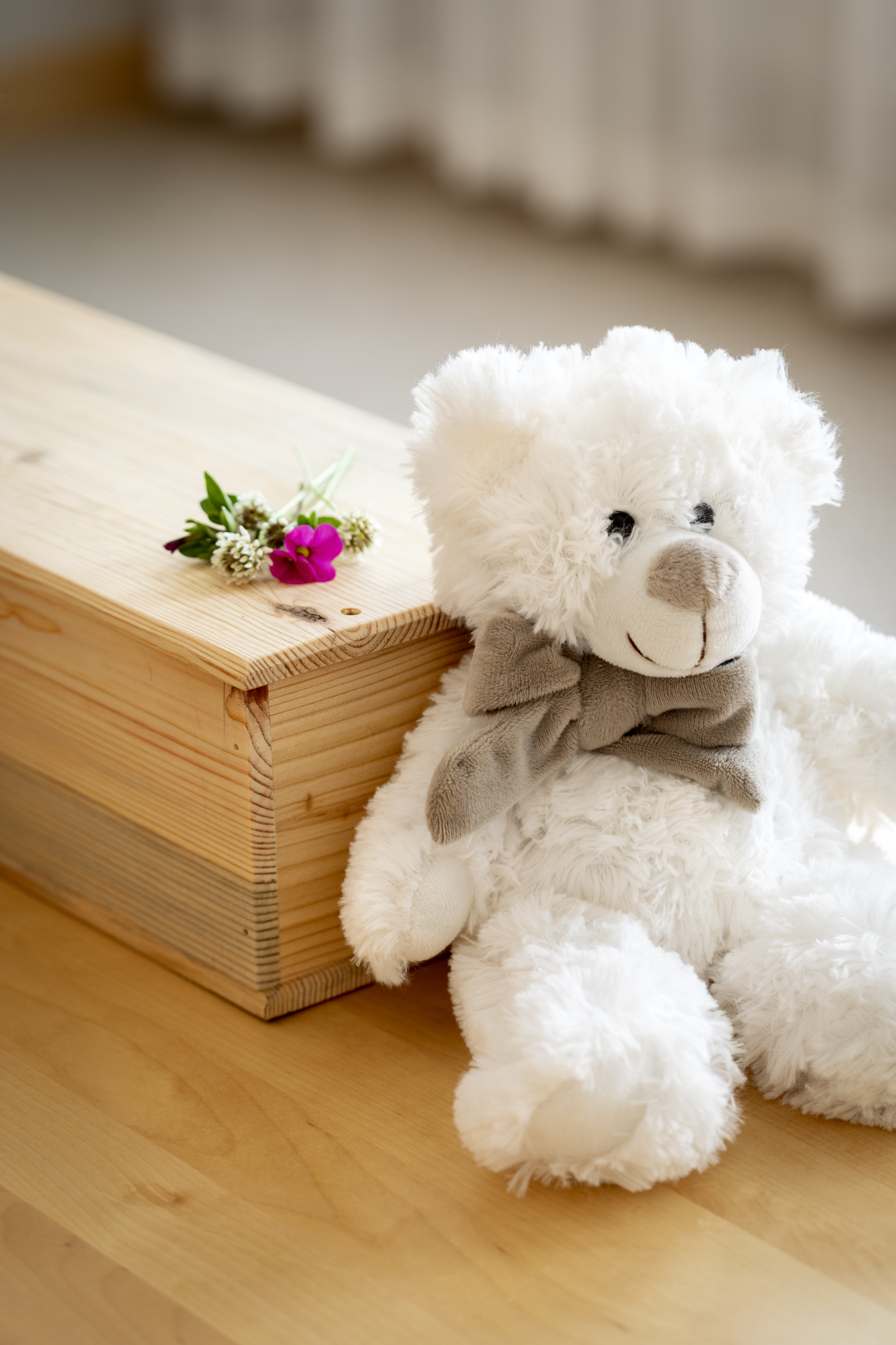 En vit nallebjörn sitter lutad mot en liten träkista. Några blommor ligger på kistan.