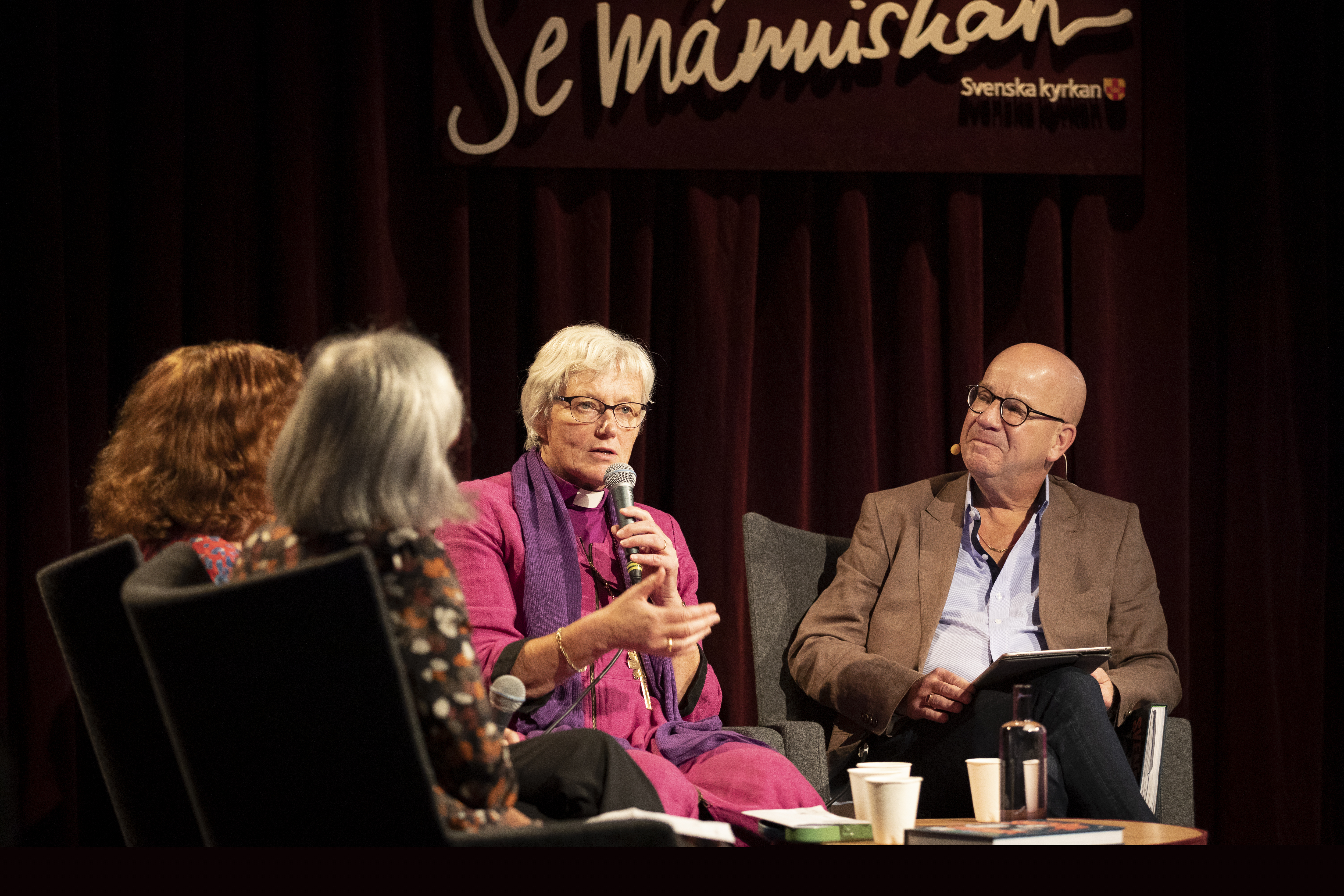 Ärkebiskop Antje Jackelén sitter i en studiomiljö och pratar i en mikrofon. På väggen bakom står det Se människan.
