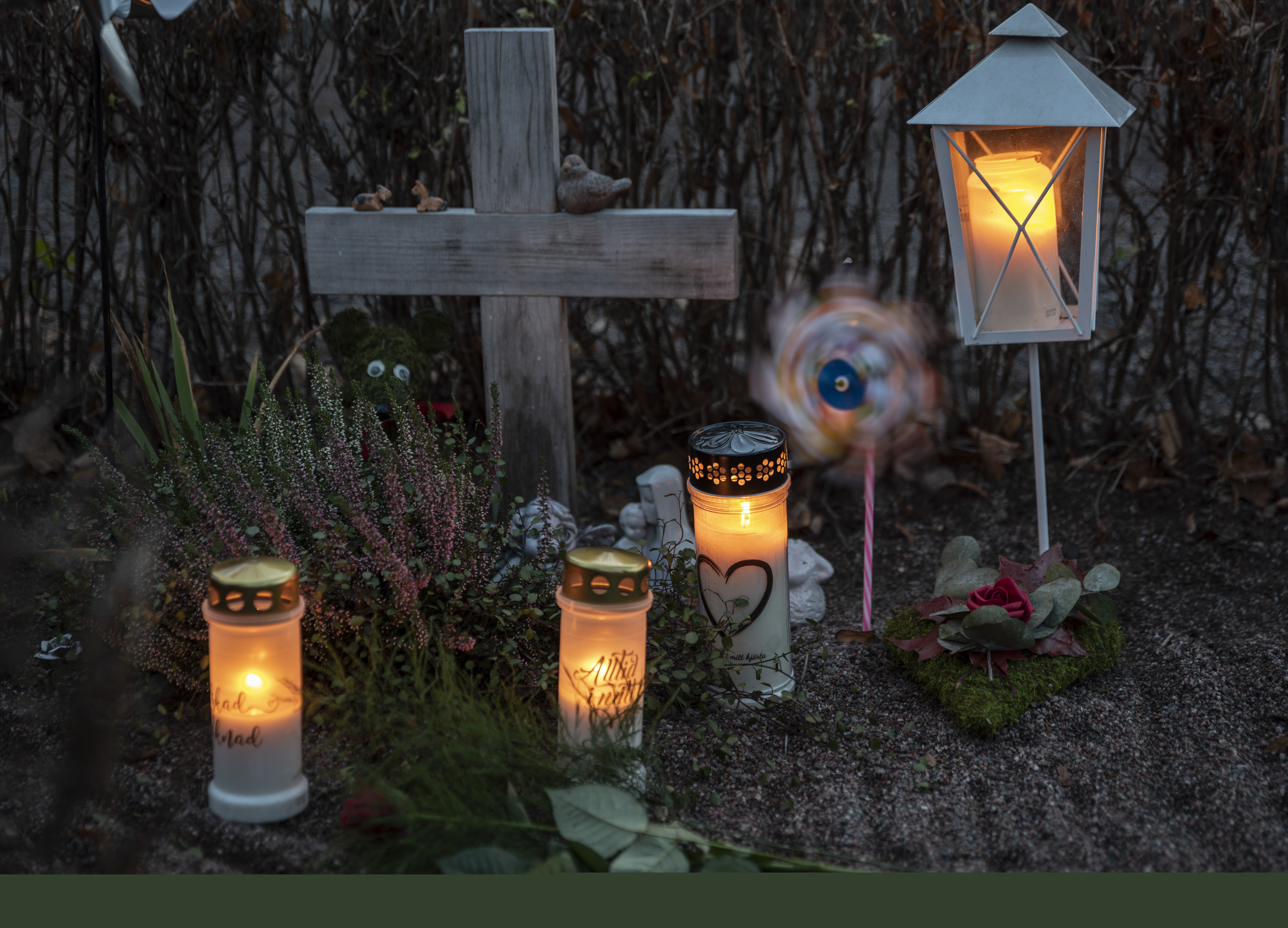 En grav med ett enkelt träkors har smyckats med gravljus, en lykta, en vindsnurra, ler- och gipsfigurer samt olika blommor.