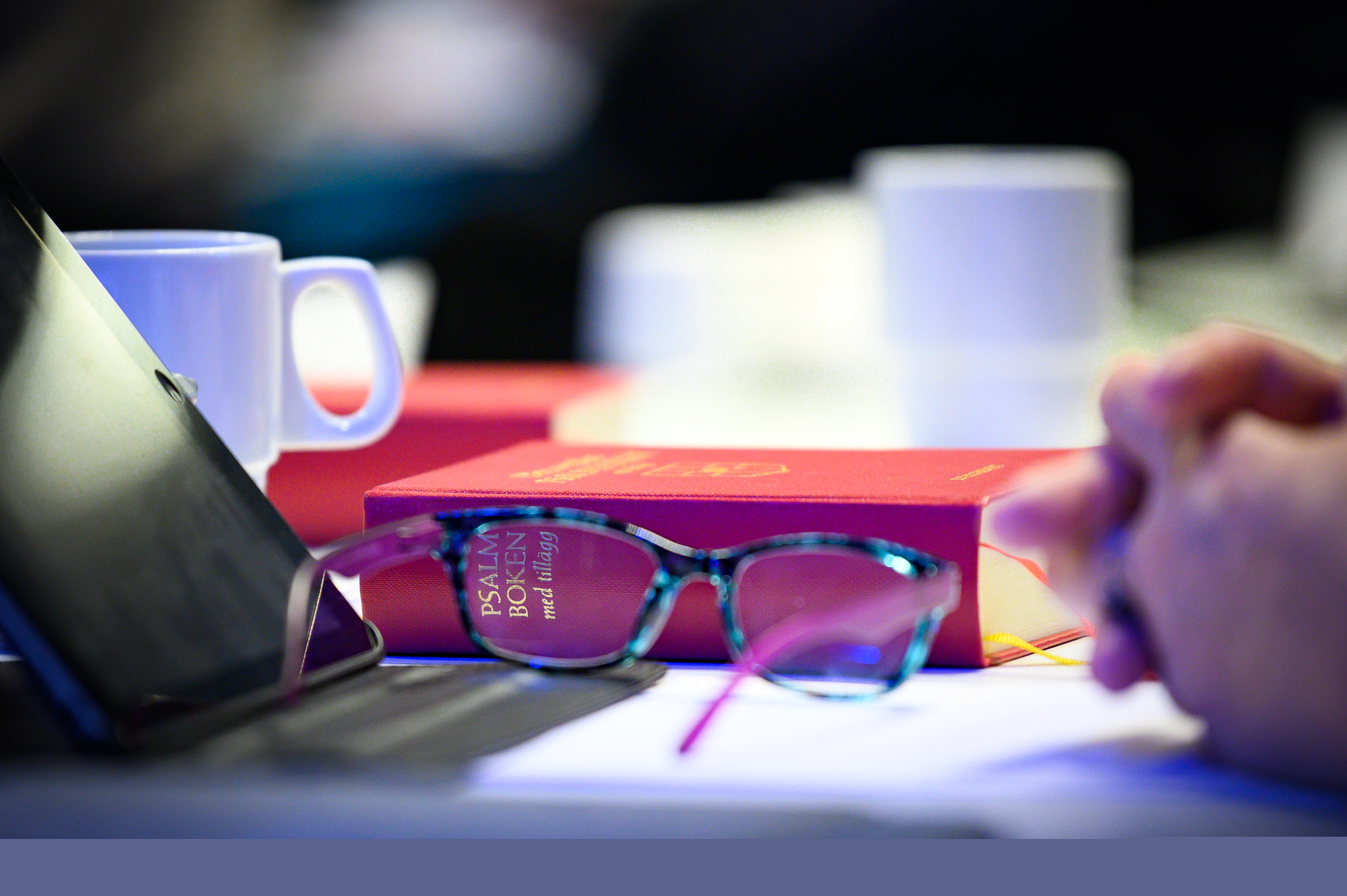 Ett par glasögon och en psalmbok ligger på bordet framför en mötesdeltagare.