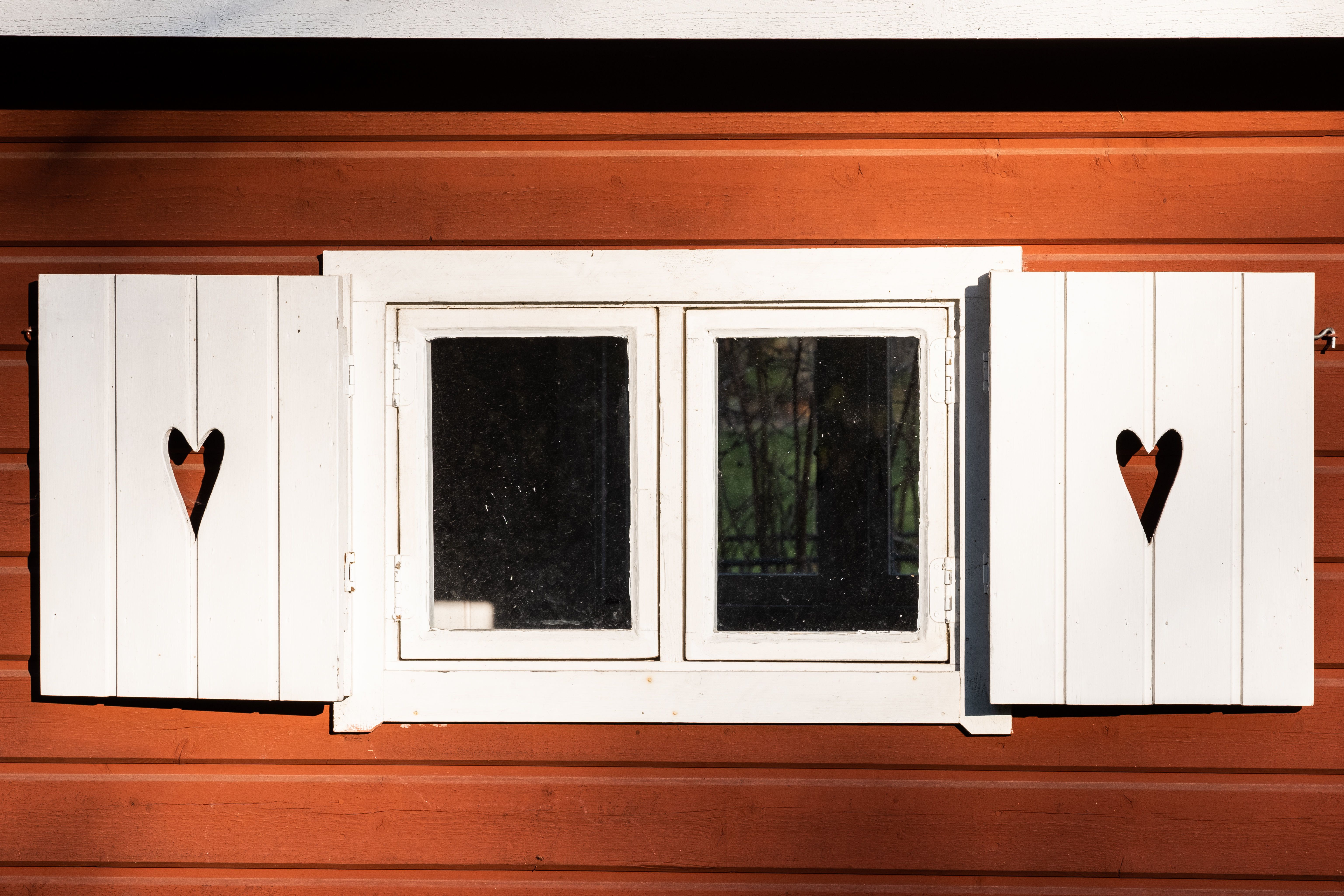 Ett fönster på ett falurött hur har fönsterluckor med urfrästa hjärtan i.