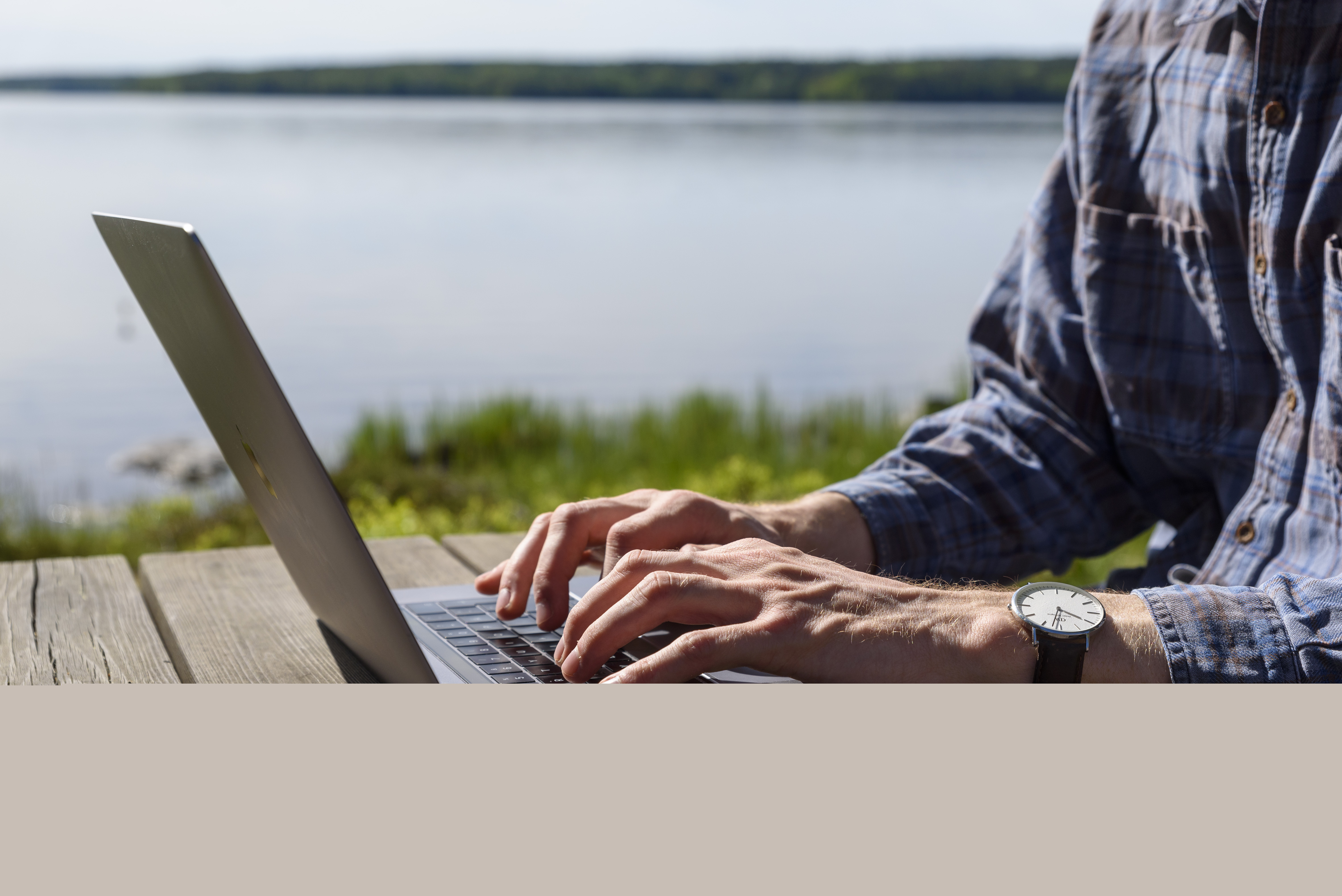 En person sitter vid ett träbord vid en sjö och jobbar på sin laptop.