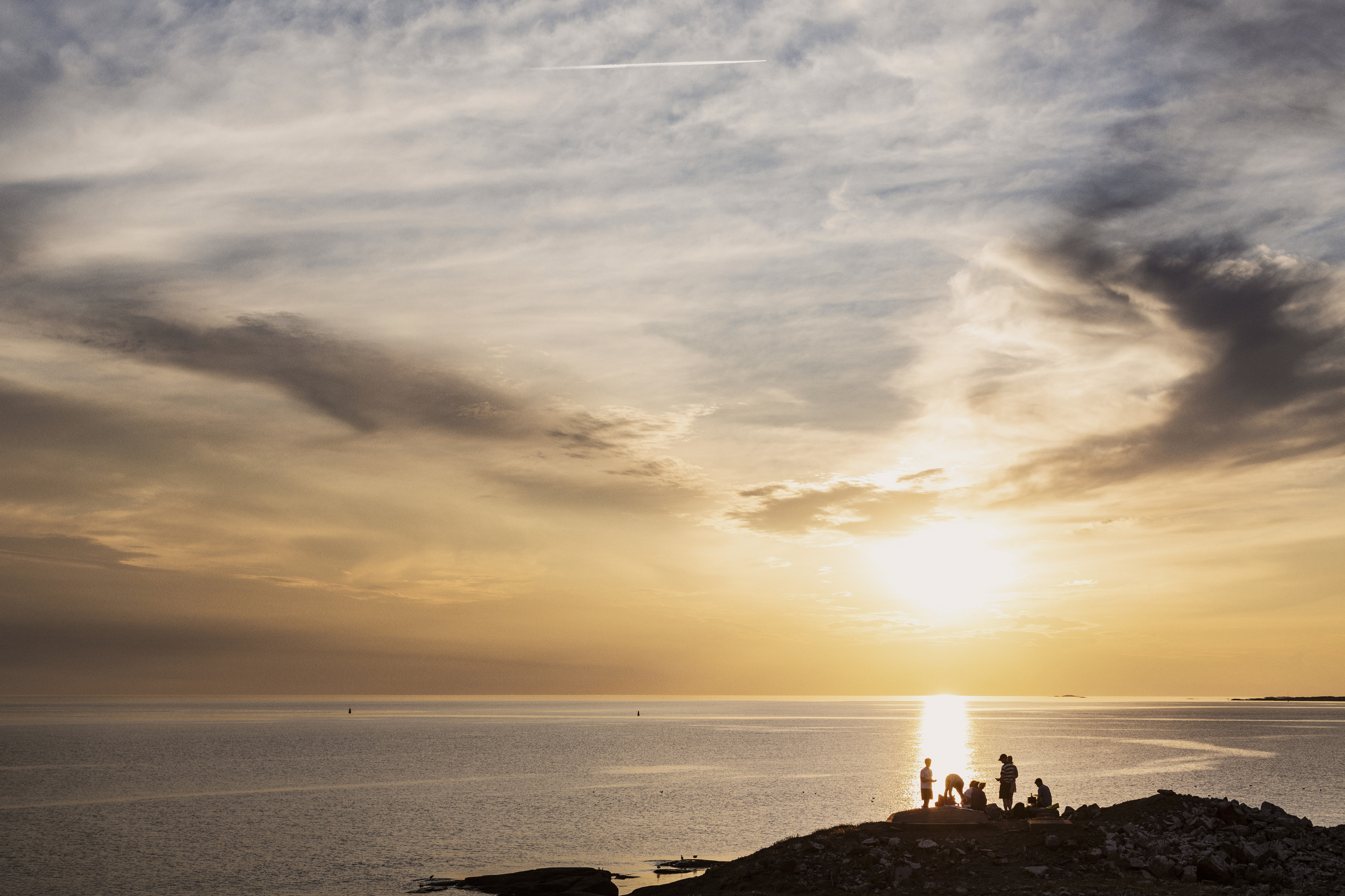 På en klippa vid havet har en grupp personer slagit sig ner för att se på solnedgången.