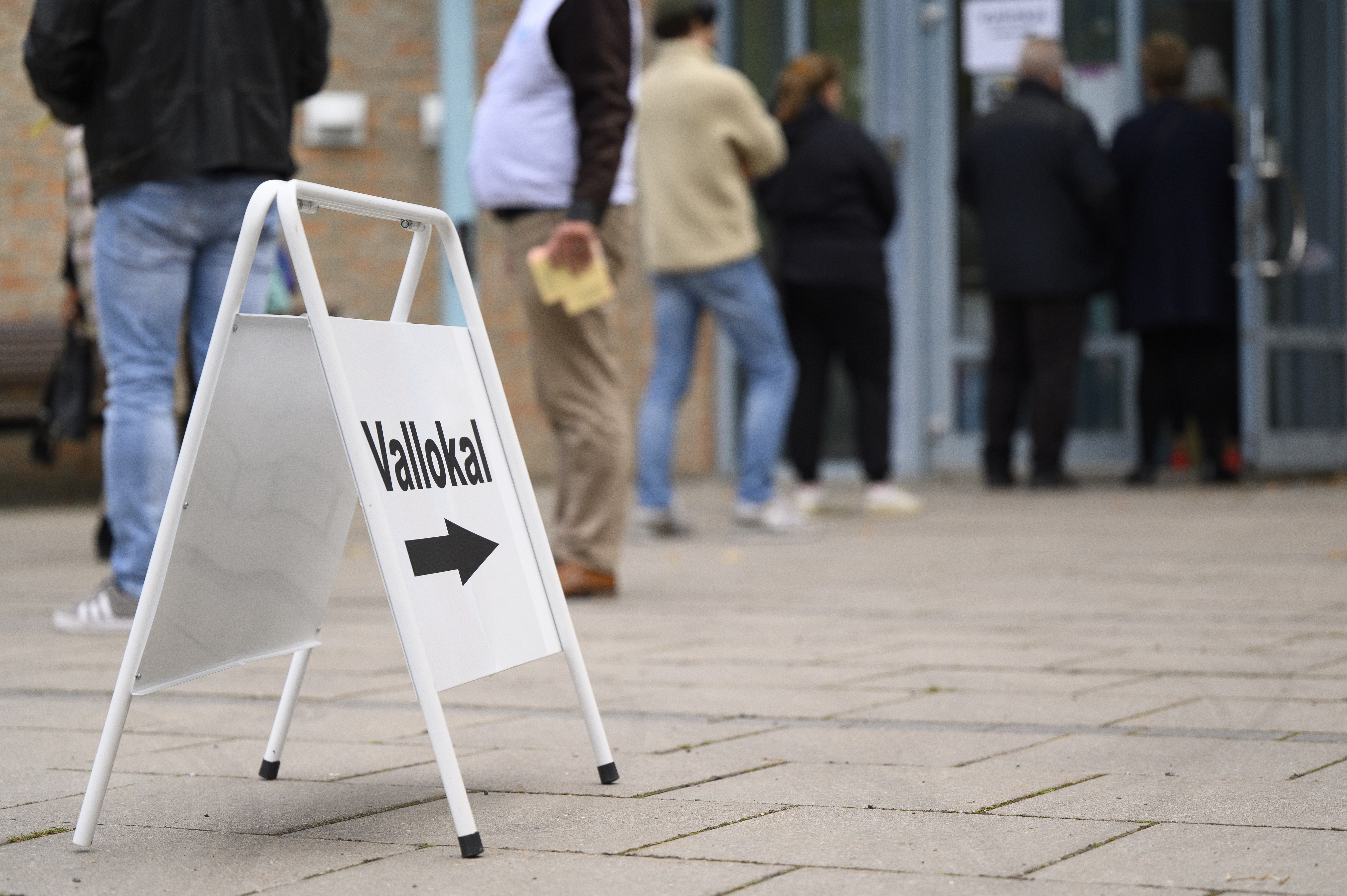 En skylt med texten Vallokal står på marken. Människor köar utanför en öppen dörr i bakgrunden.