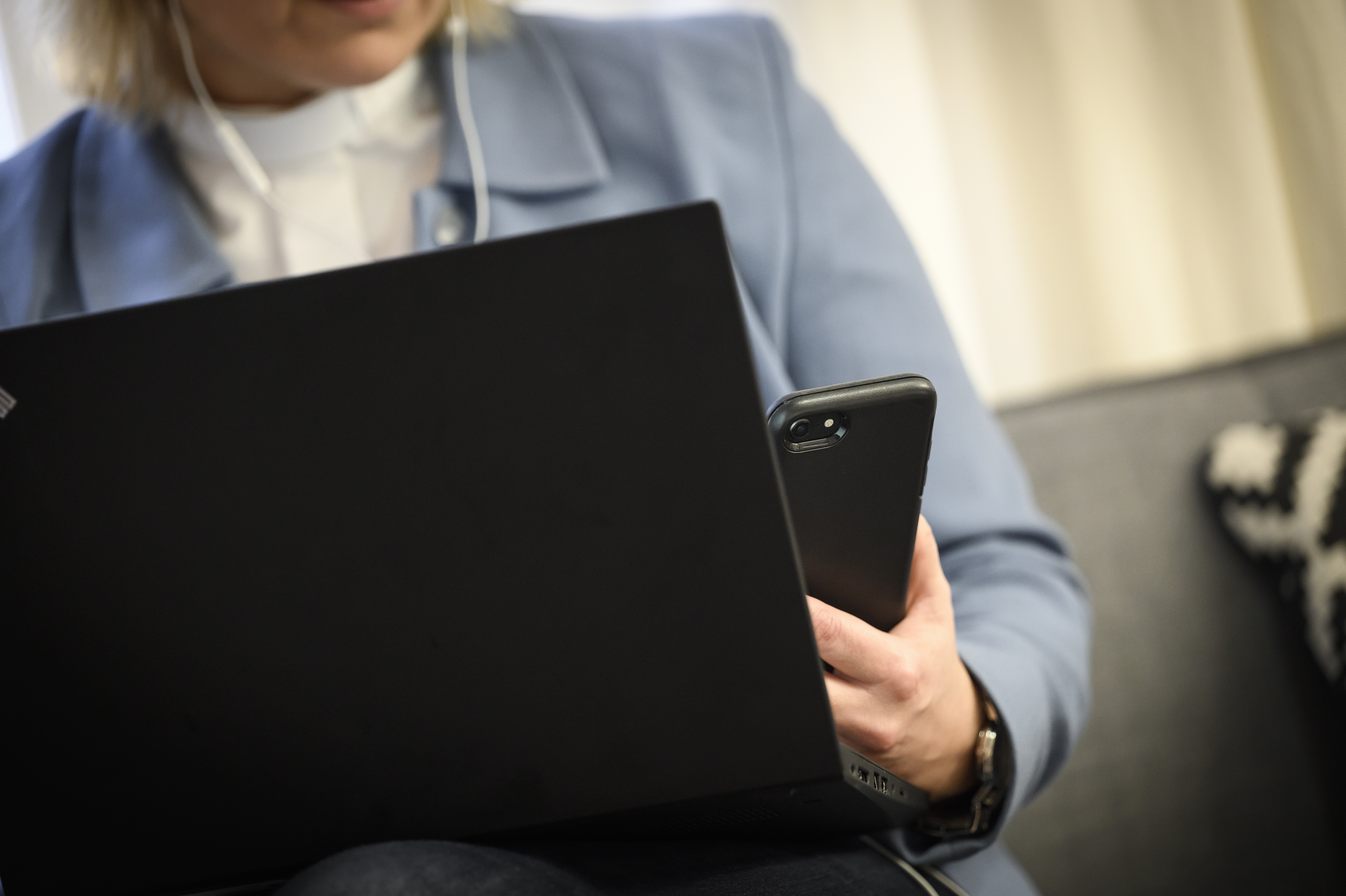 En kvinnlig präst i ljusblå kavaj sitter i en soffa med en laptop i knät och en mobiltelefon i handen.