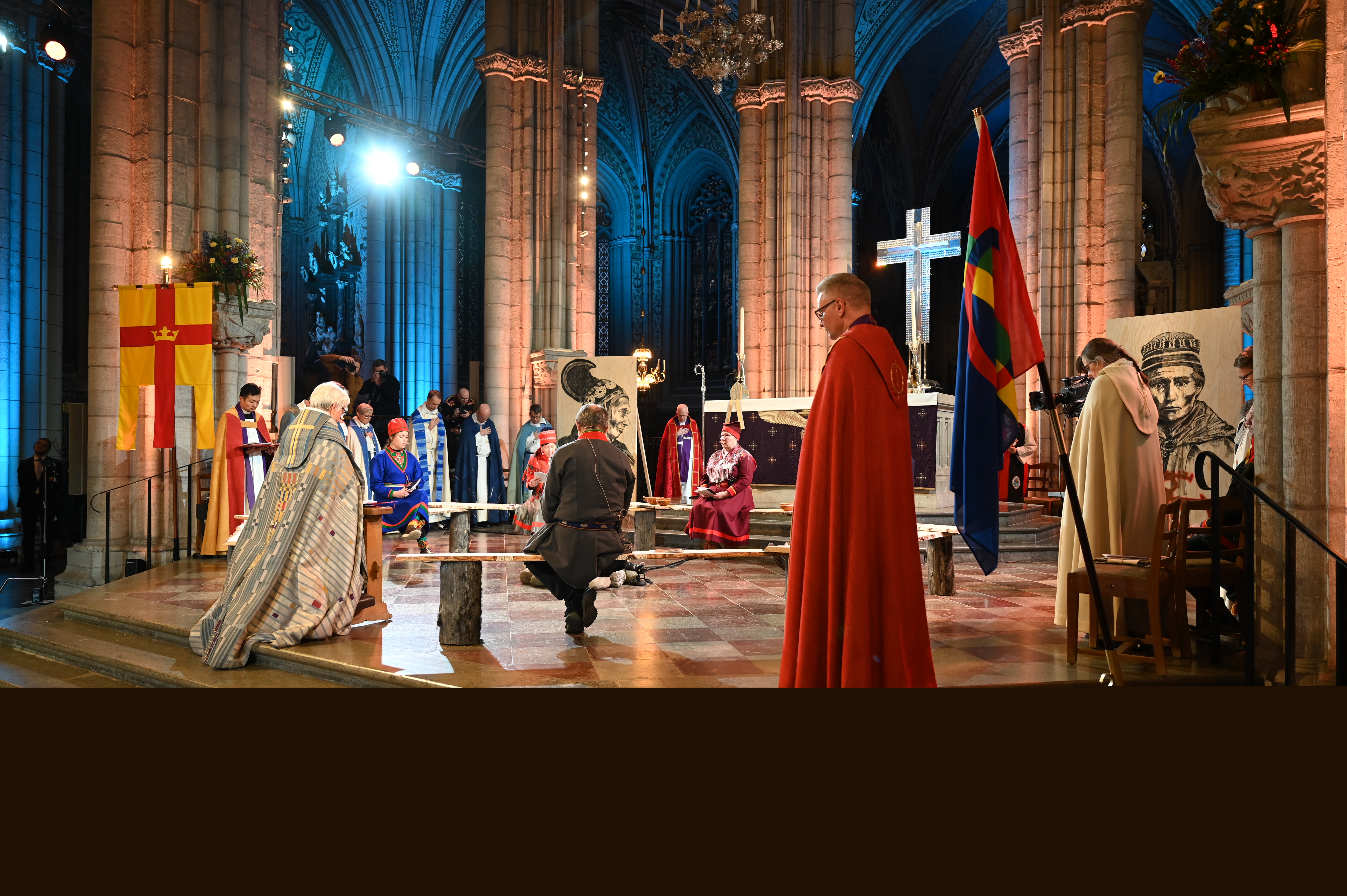 Fyra personer i samedräkt sitter på bänkar runt en lägereld framför Uppsala domkyrkas altare. Ärkebiskop Antje Jackelén knäböjer.
