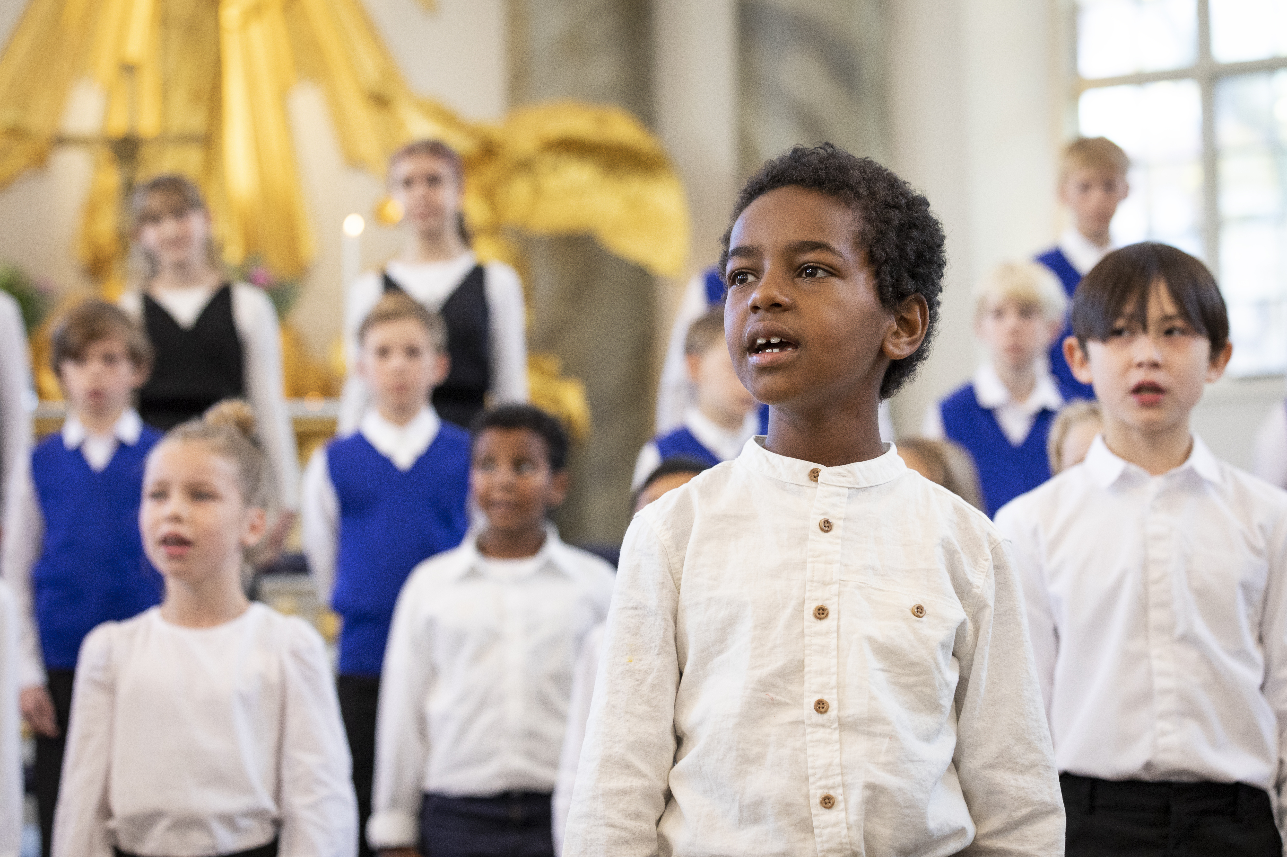 Medlemmar i en barnkör står och sjunger i en kyrka. En pojke står framför de andra och sjunger.