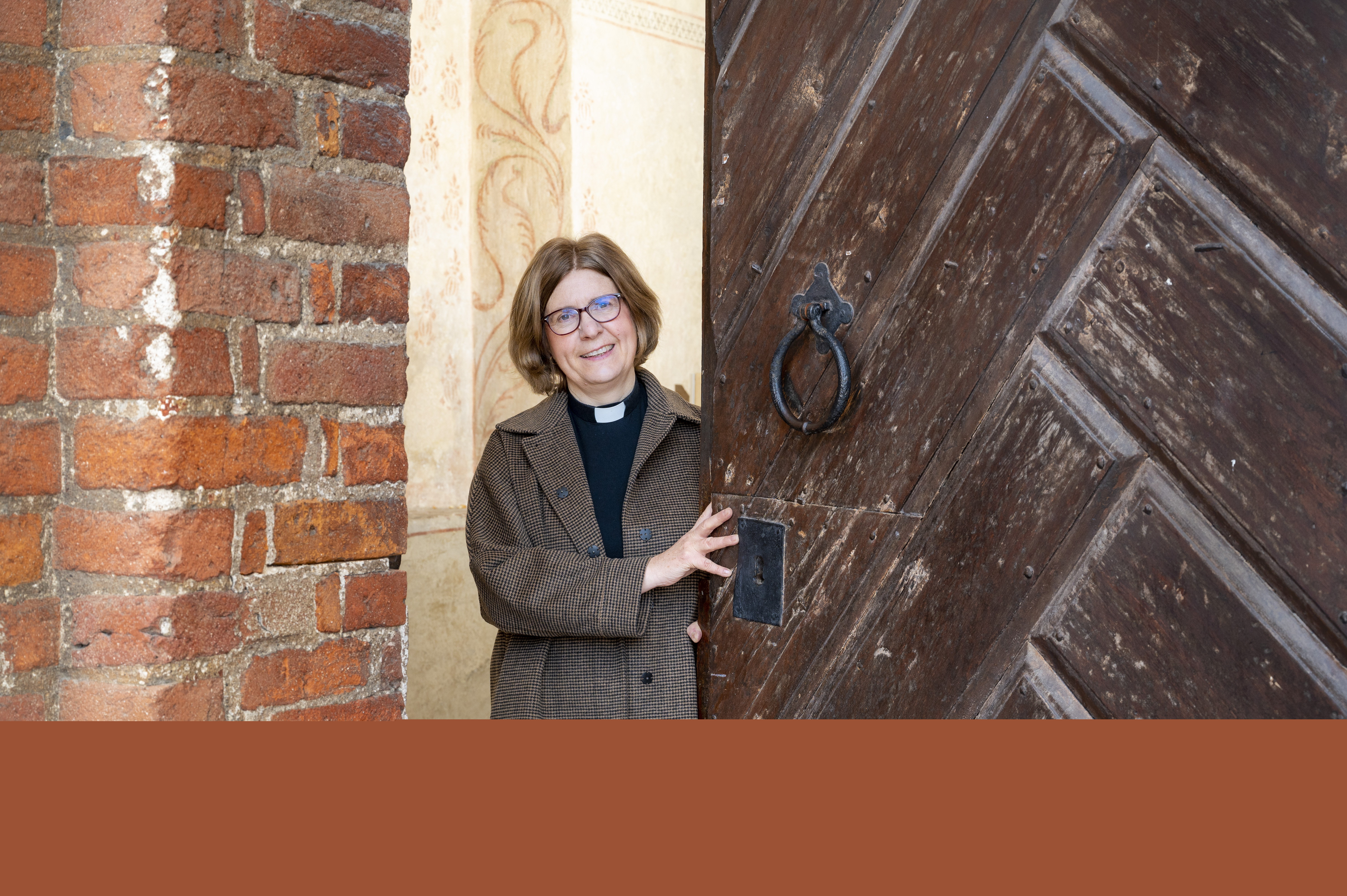En kvinnlig präst står i dörröppningen till en tegelkyrka.