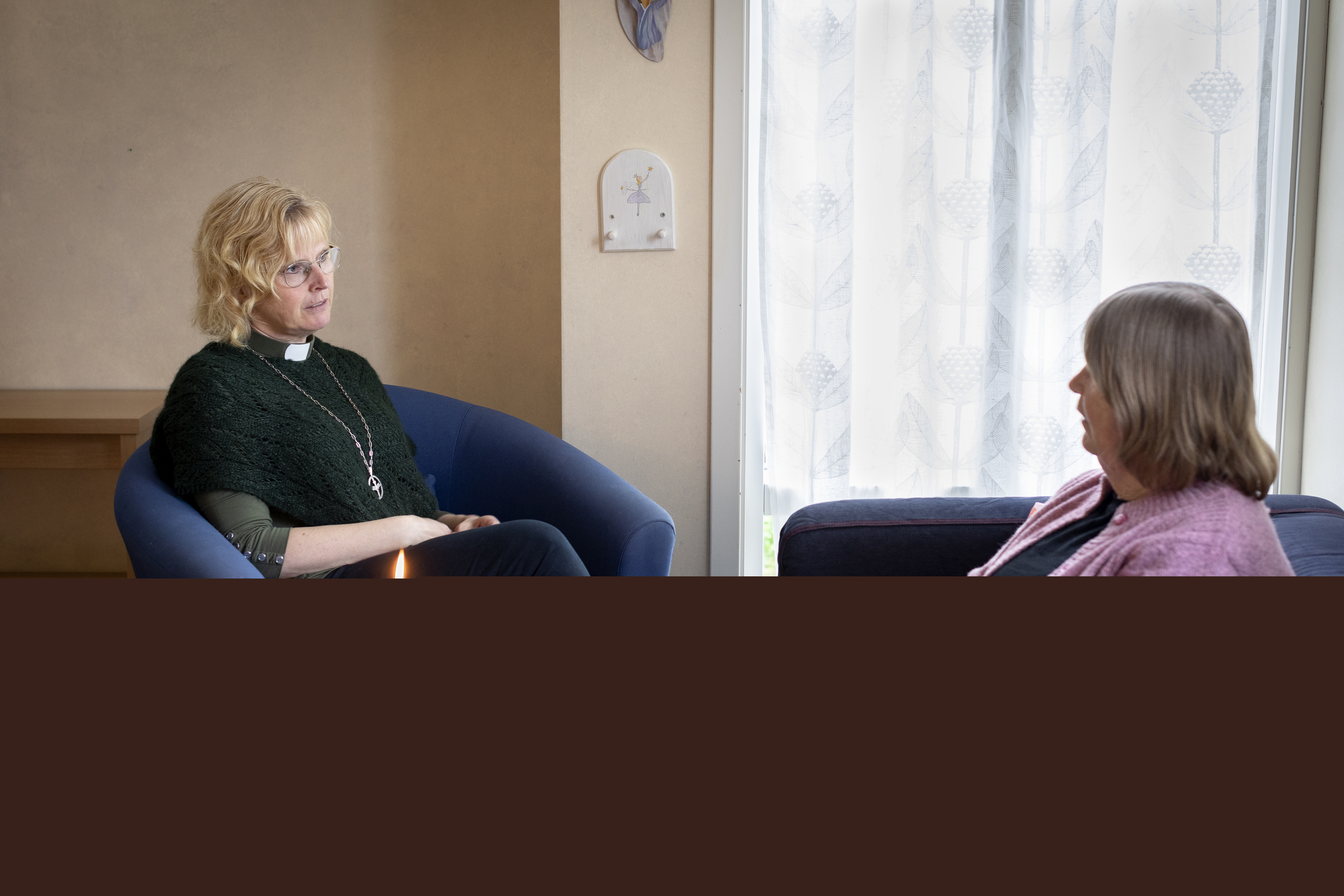 En kvinnlig diakon med grön frimärksskjorta och diakonemblem om halsen sitter i en fåtölj och samtalar med en annan kvinna.
