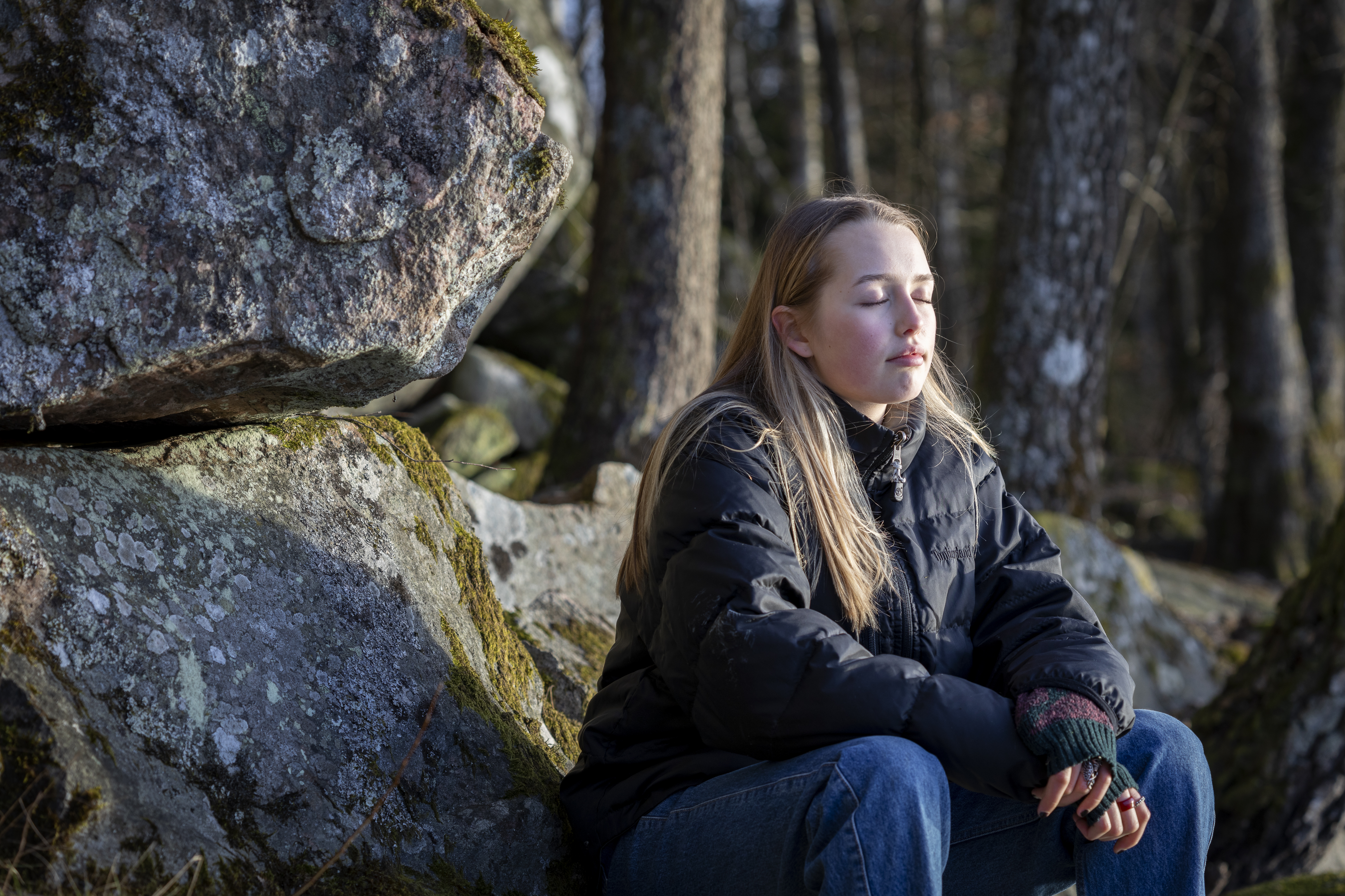 En tonårstjej sitter på en sten i skogen. Hon har ansiktet vänt mot solen och ögonen slutna.