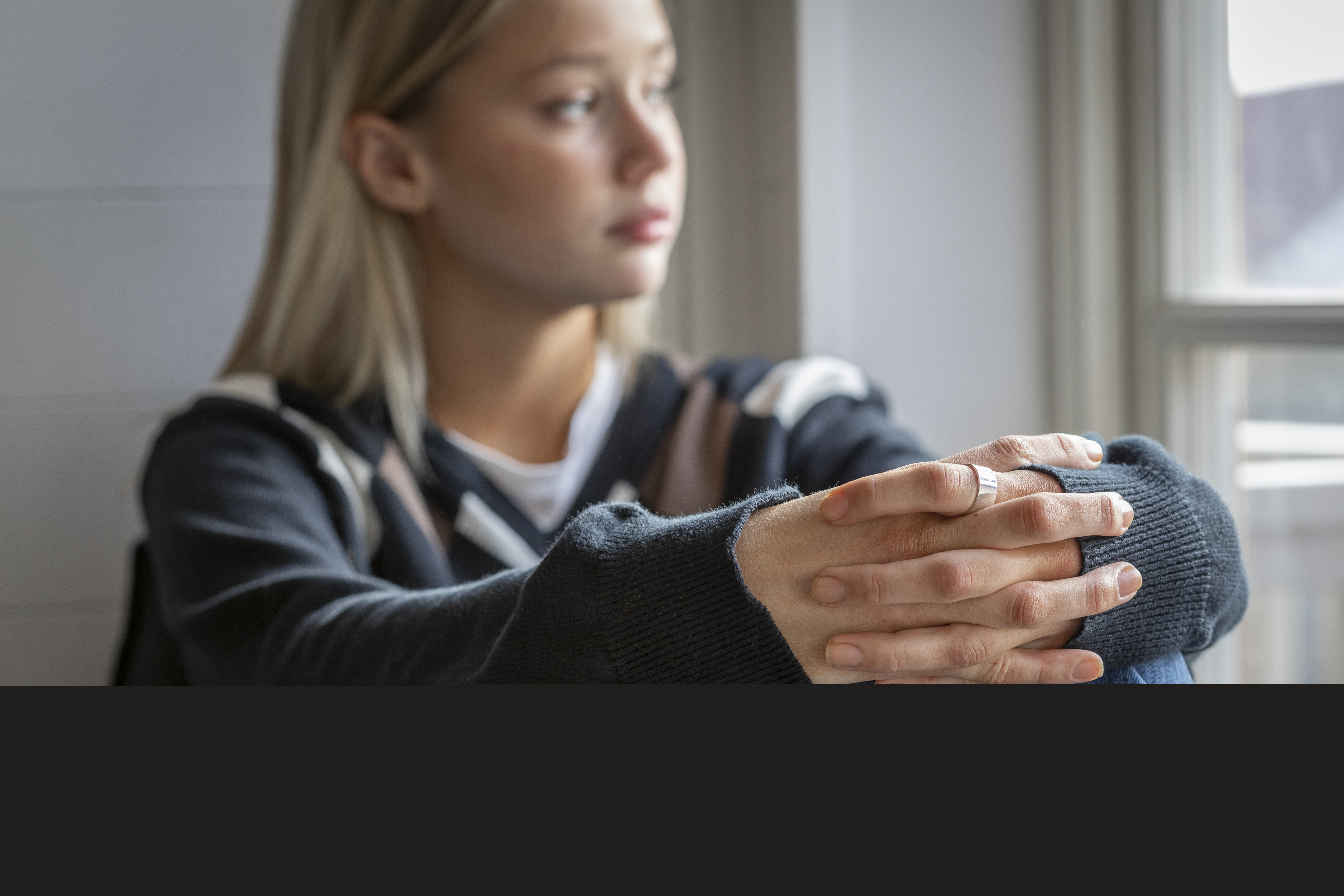 En tonårstjej sitter vid ett fönster med händerna om knäna.