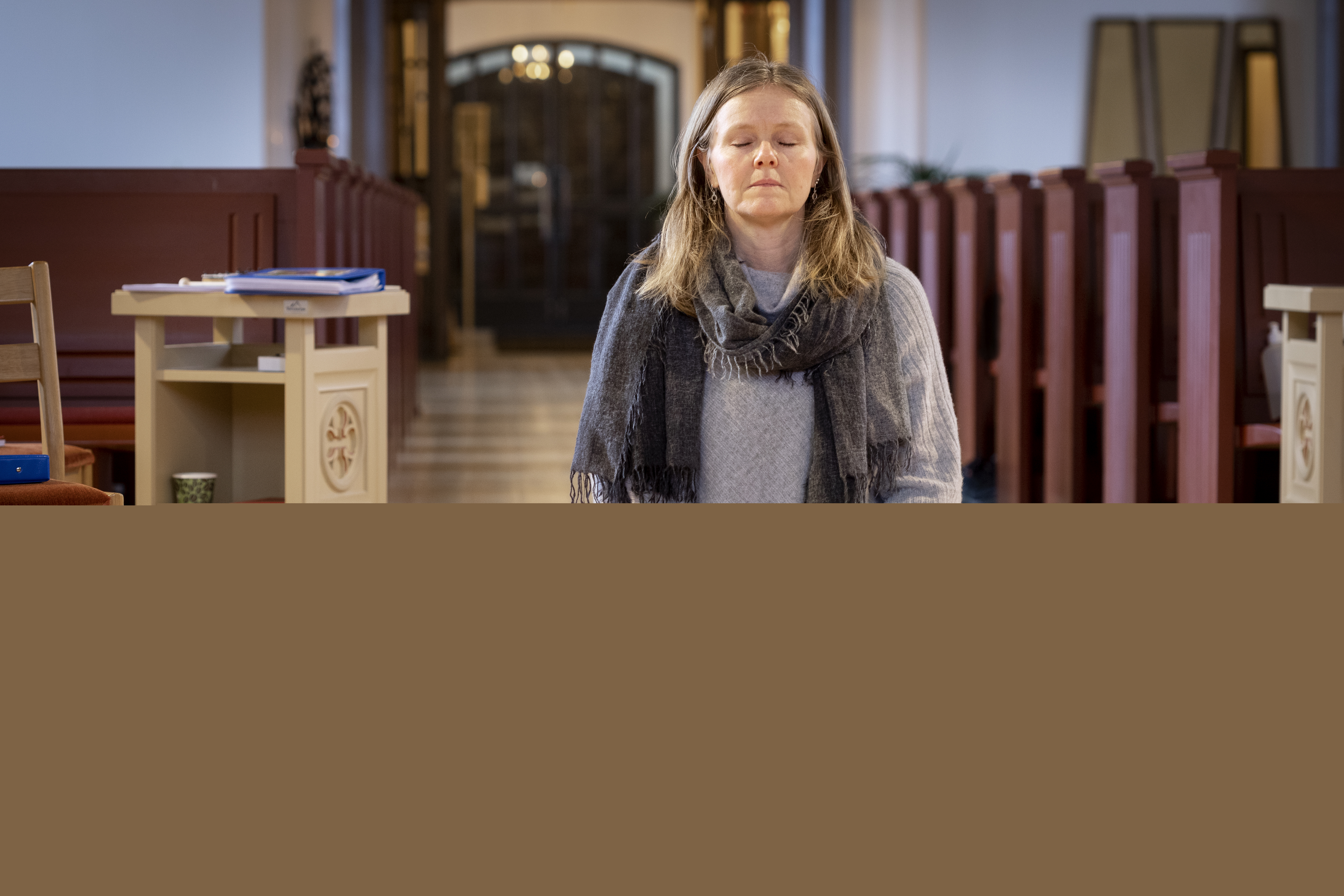 En kvinna sitter på en bönepall på en kyrkas golv, med knäppta händer och slutna ögon.