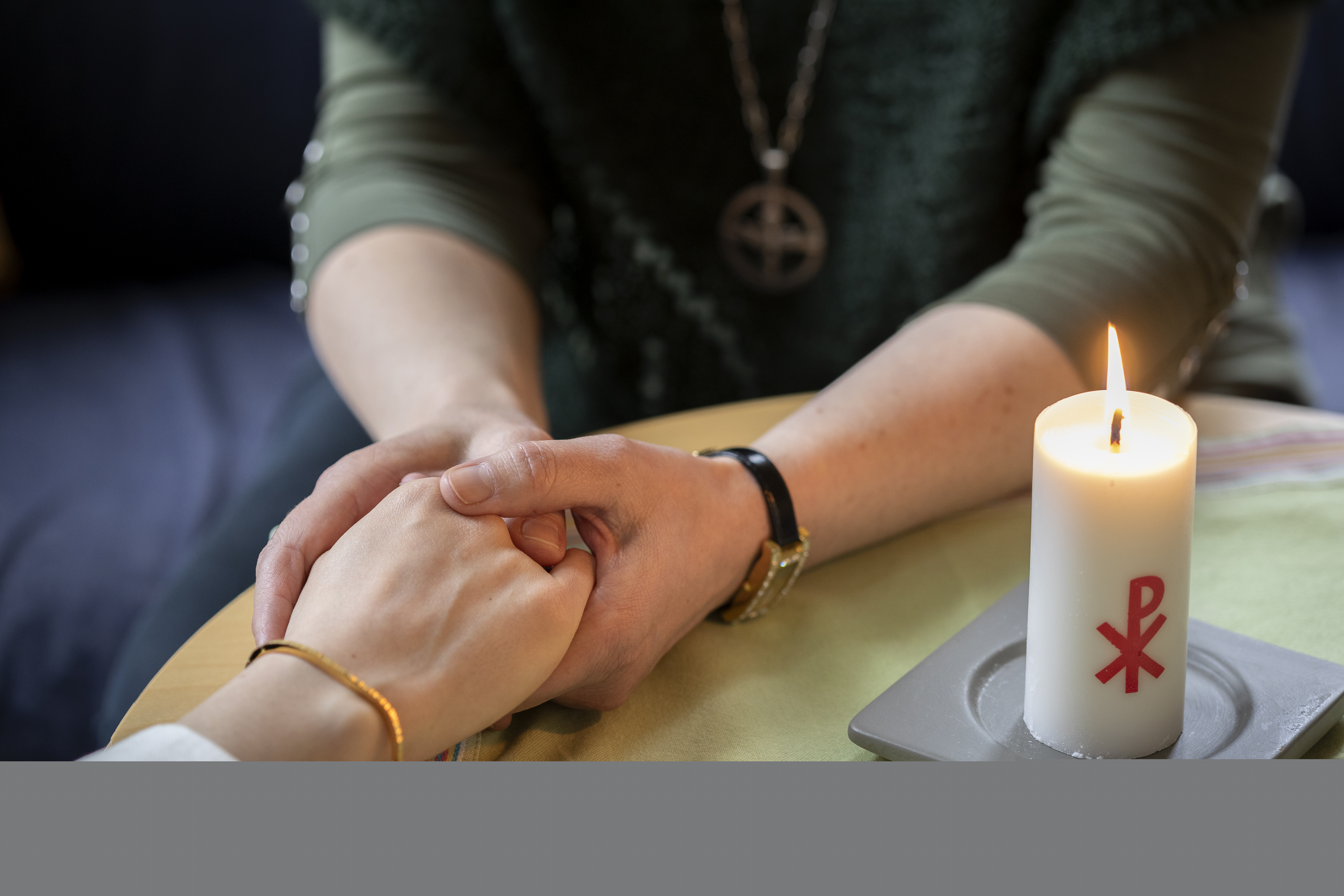 En diakon håller en annans hand i sina händer. Ett stearinljus med kristusmonogram brinner på bordet mellan dem.