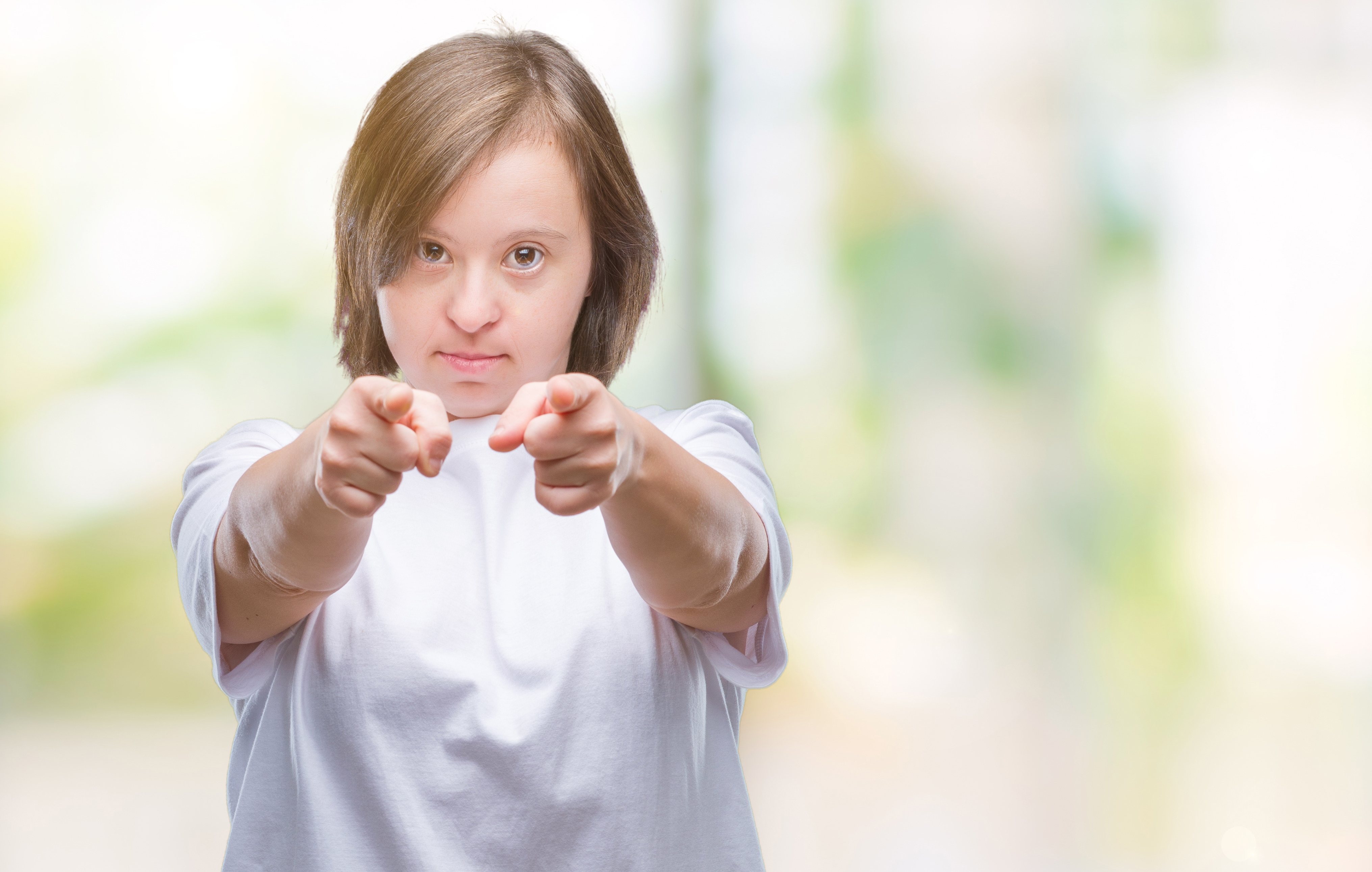 Ung kvinna med Downs syndrom pekar med båda sina pekfingrar mot kameran. Hon ar en vit kortärmad t-shirt. Hon riktar sin blick tydligt mot kameran
