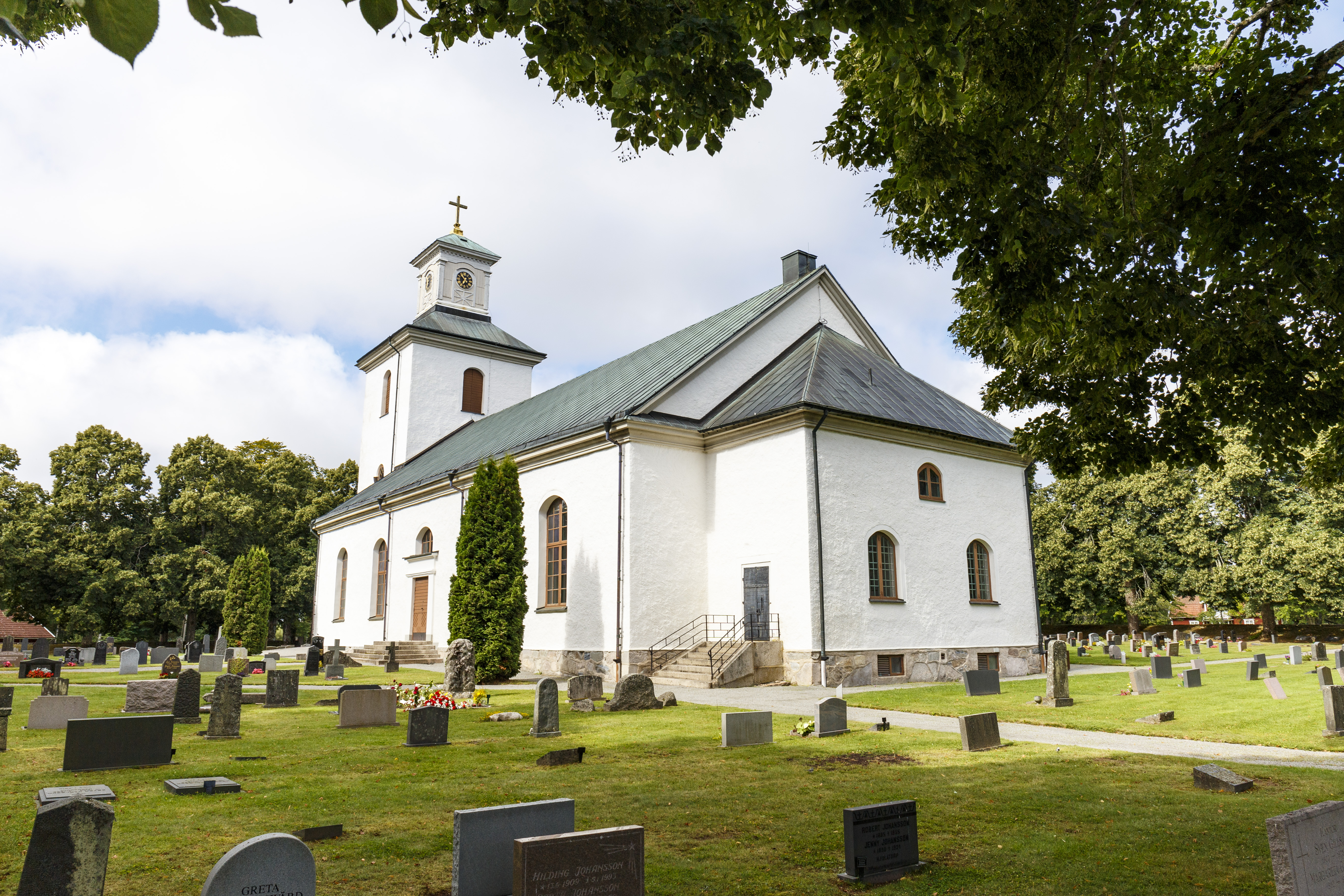 En vitputsad stenkyrka med ärgat tak. Gravstenar och grönskande träd i omgivningen.