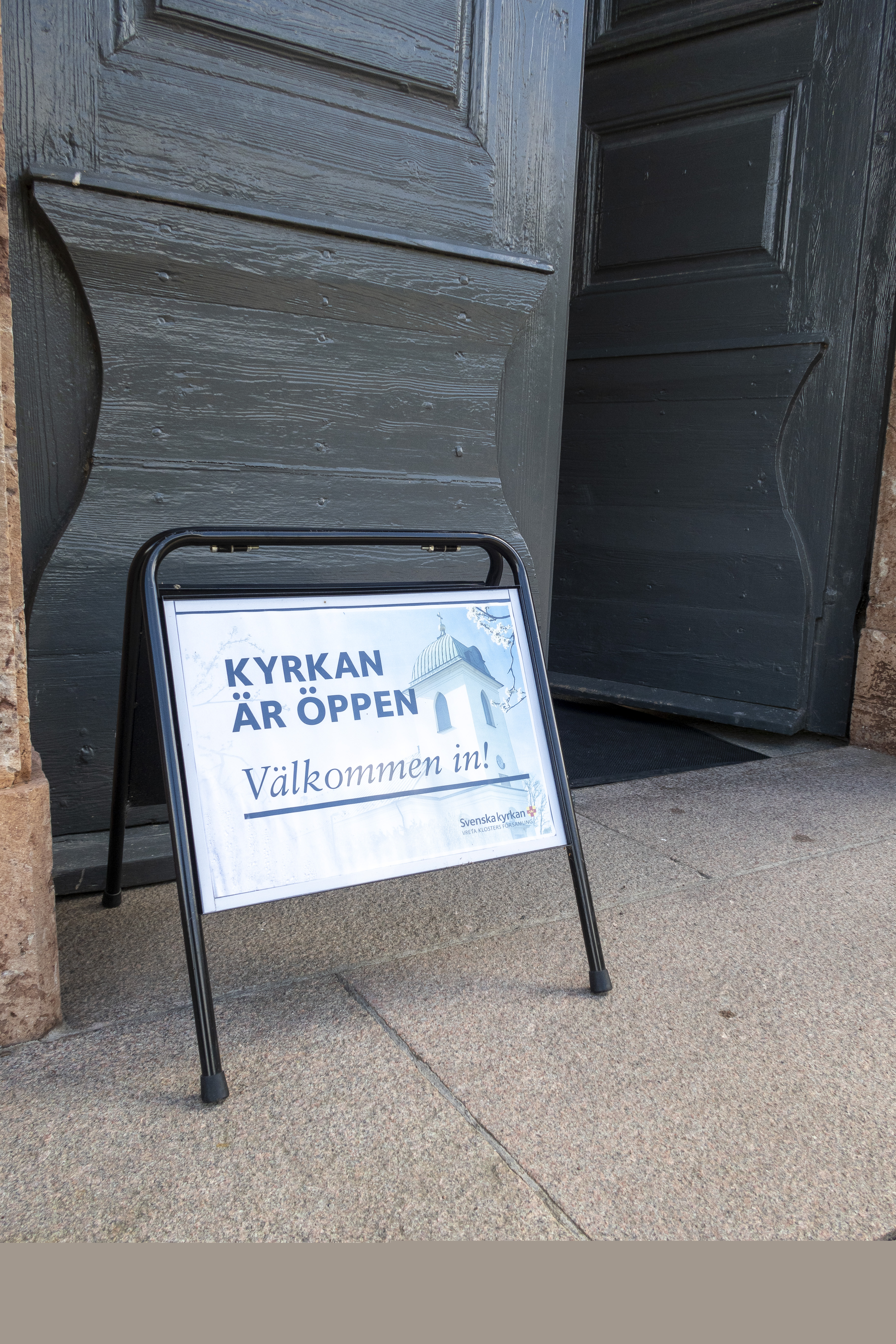 En skylt med texten Kyrkan är öppen välkommen in, står utanför en port.