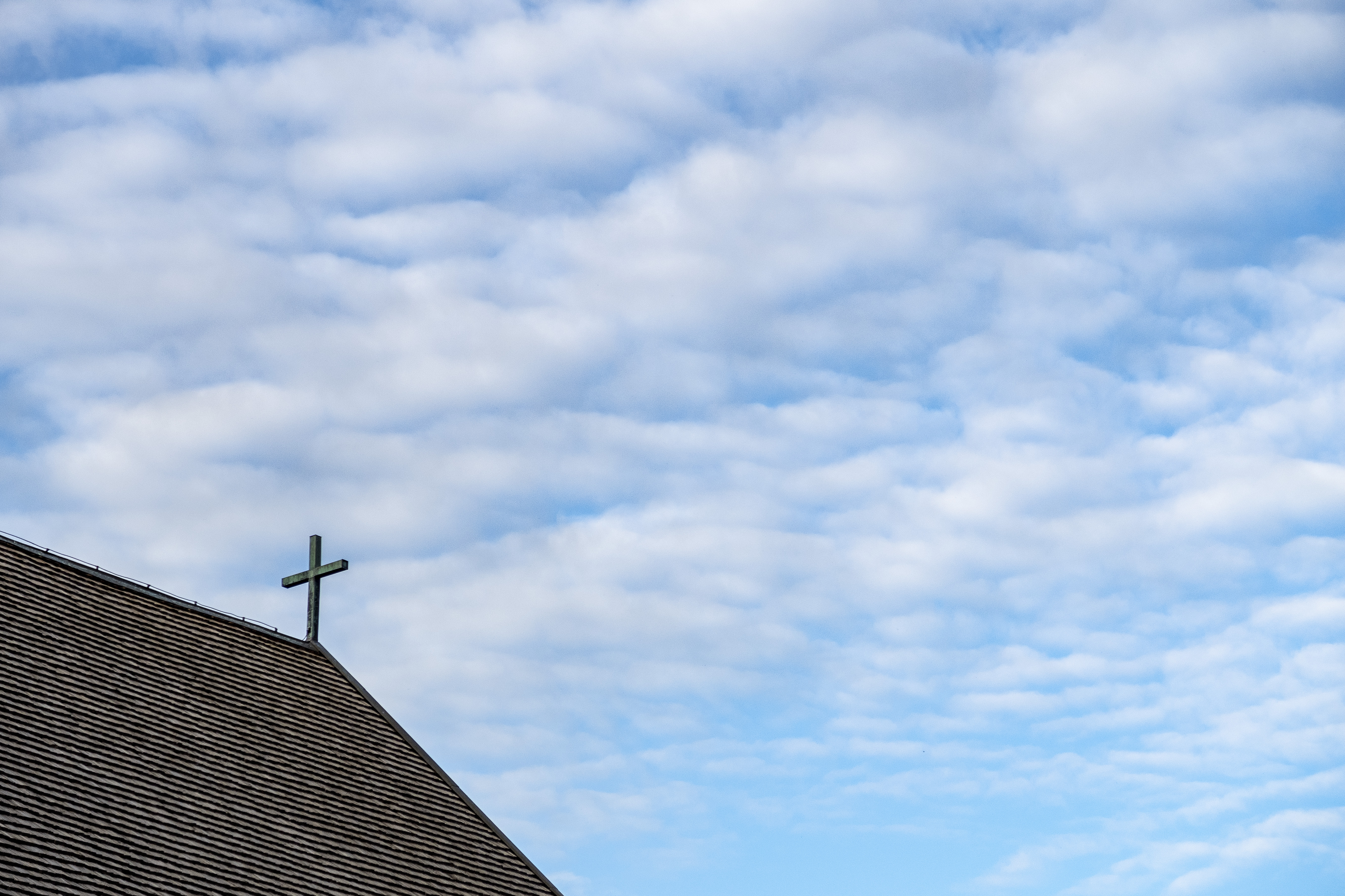Ett kyrktak med ett litet kors mot en blå och molnig himmel.