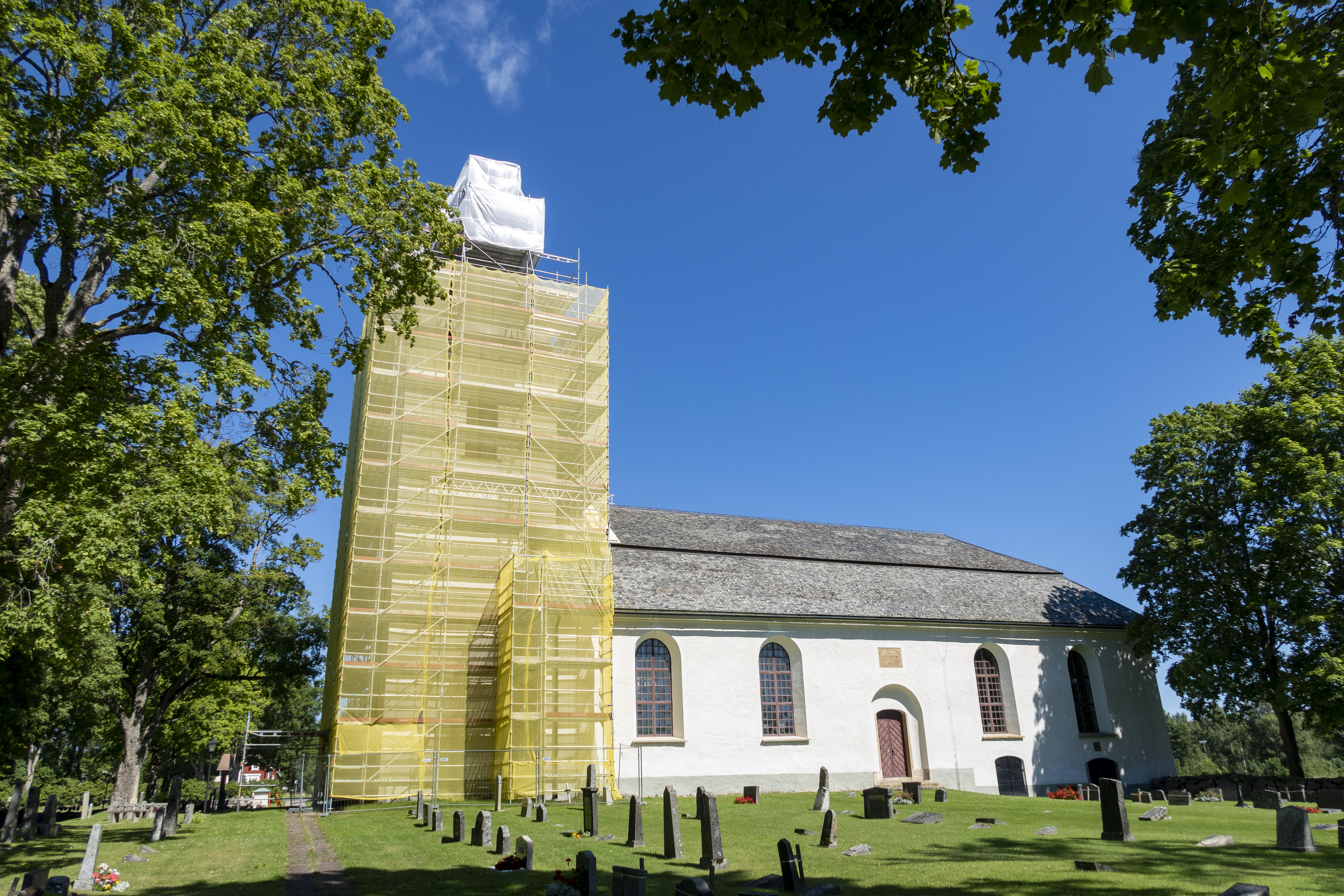 Byggnadsställningar runt ett kyrktorn. En kyrkogård framför den vita kyrkan.