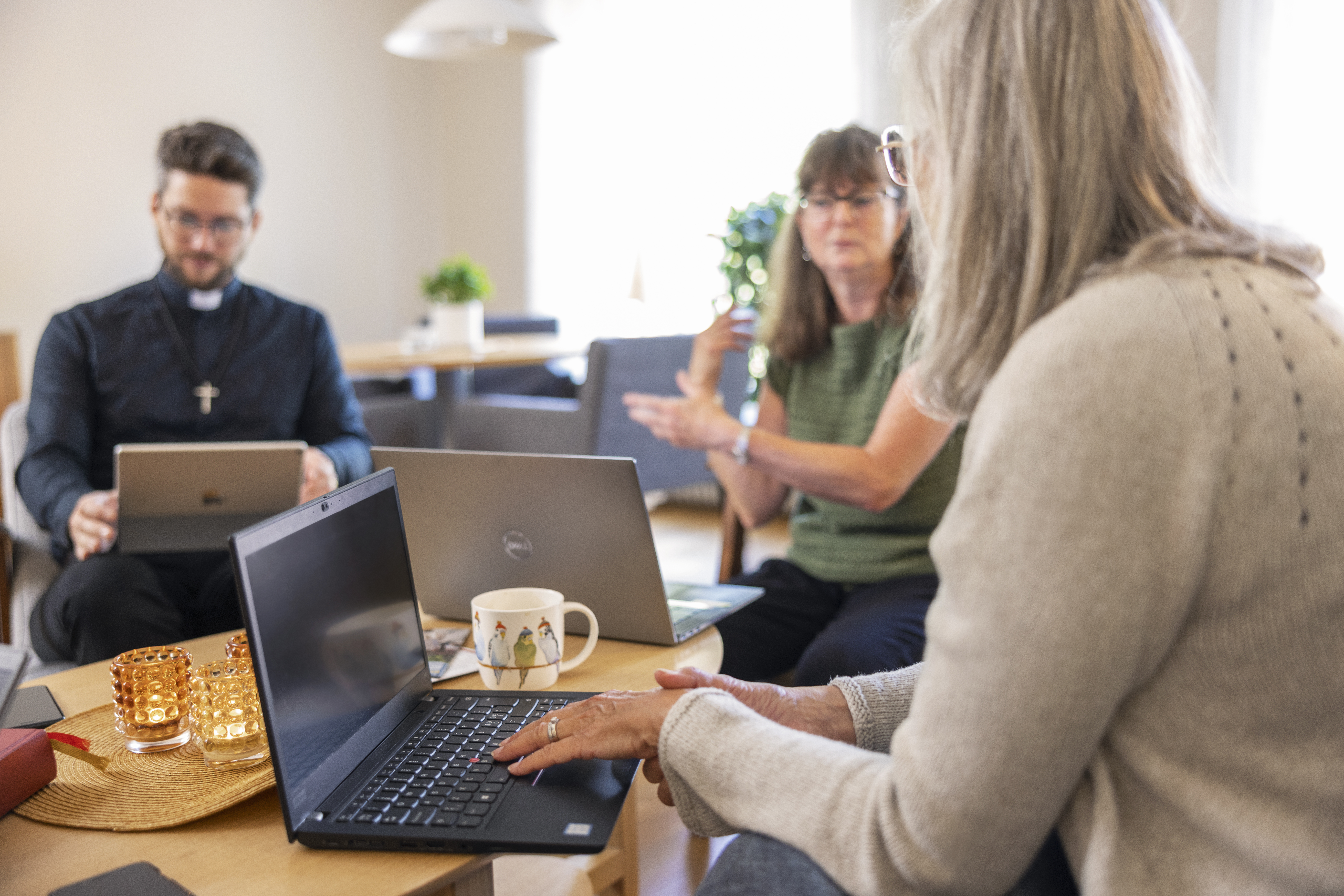 En manlig präst och två kvinnor sitter vid ett bord med varsin laptop och samtalar.