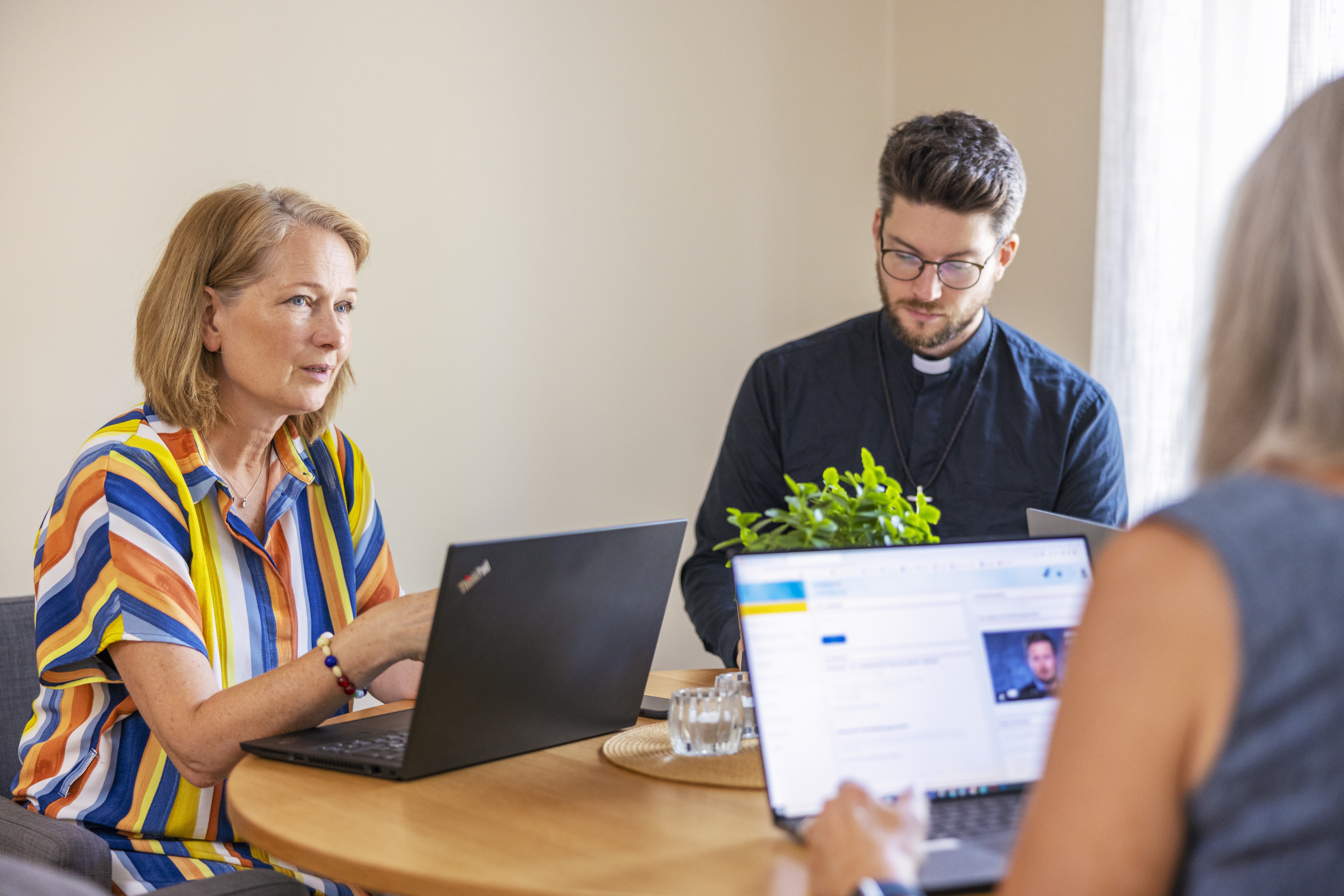 En manlig präst och några andra personer sitter runt ett bord med varsin laptop och samtalar.