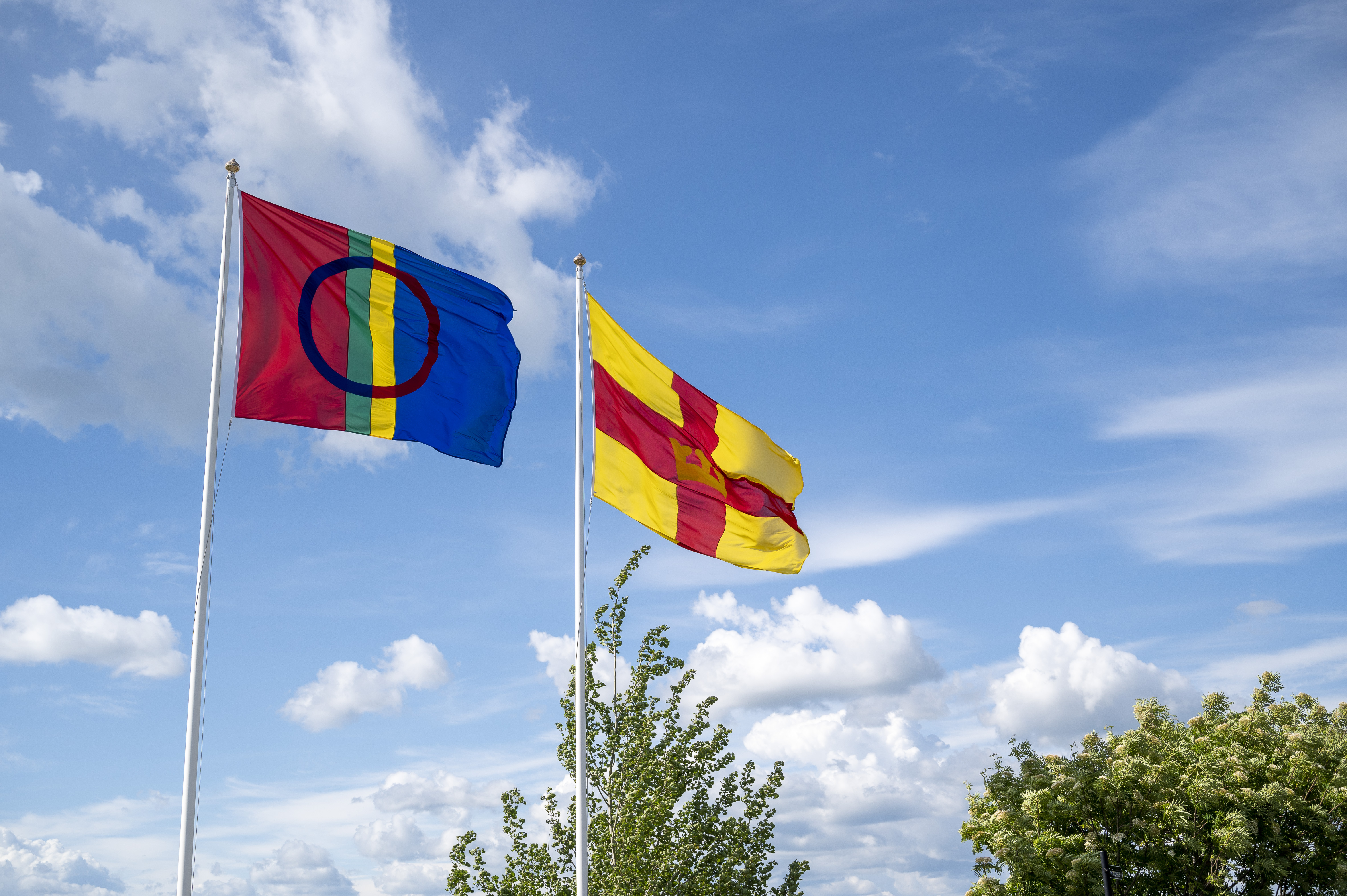 Två flaggstänger med den samiska flaggan och Svenska kyrkans flagga mot en blå himmel.
