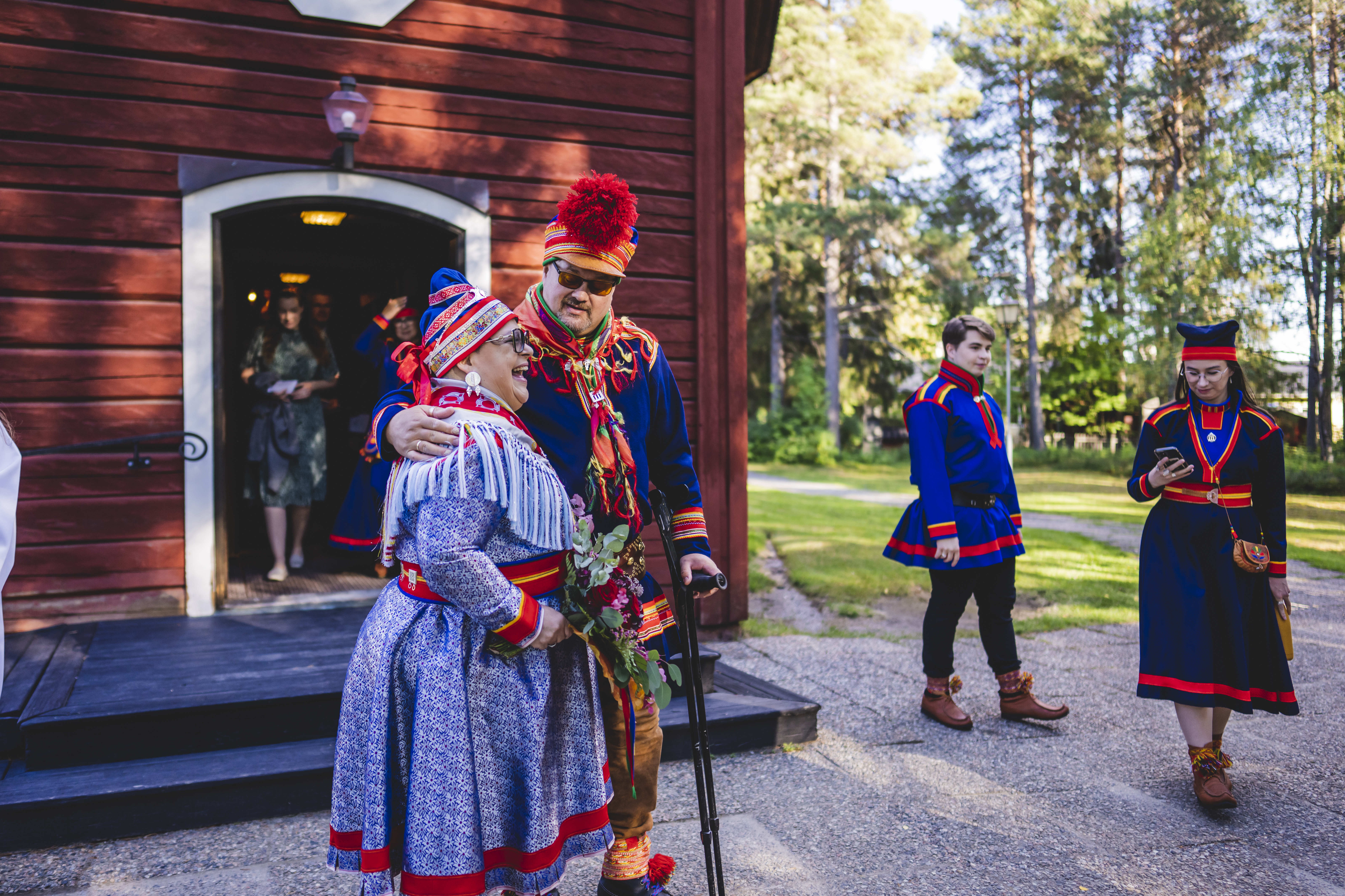 Några personer i samiska folkdräkter står utanför en röd kyrka i trä.