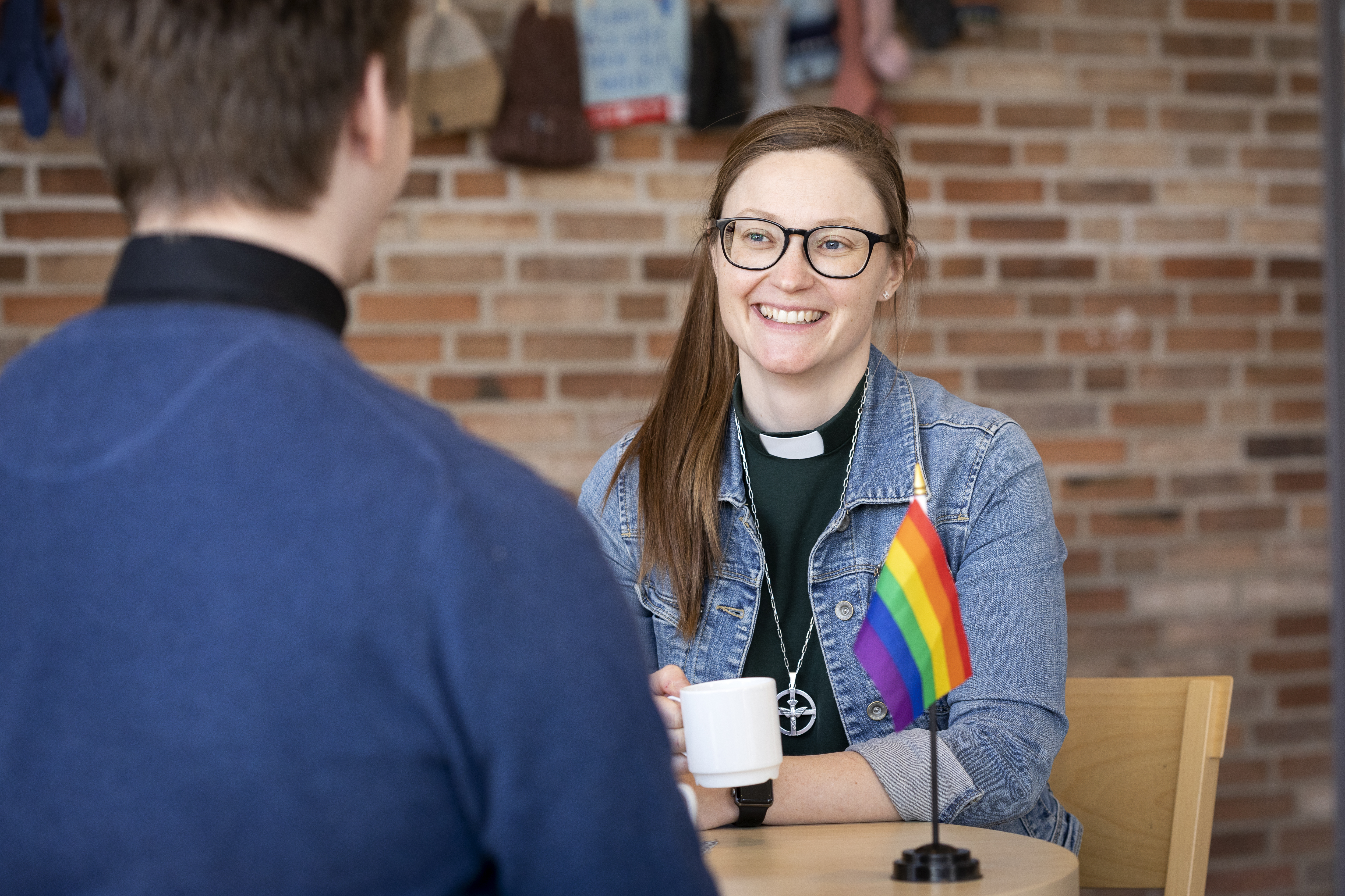 En kvinnlig diakon sitter på ett fik och samtalar med en kille. En regnbågs-flagga står på bordet.