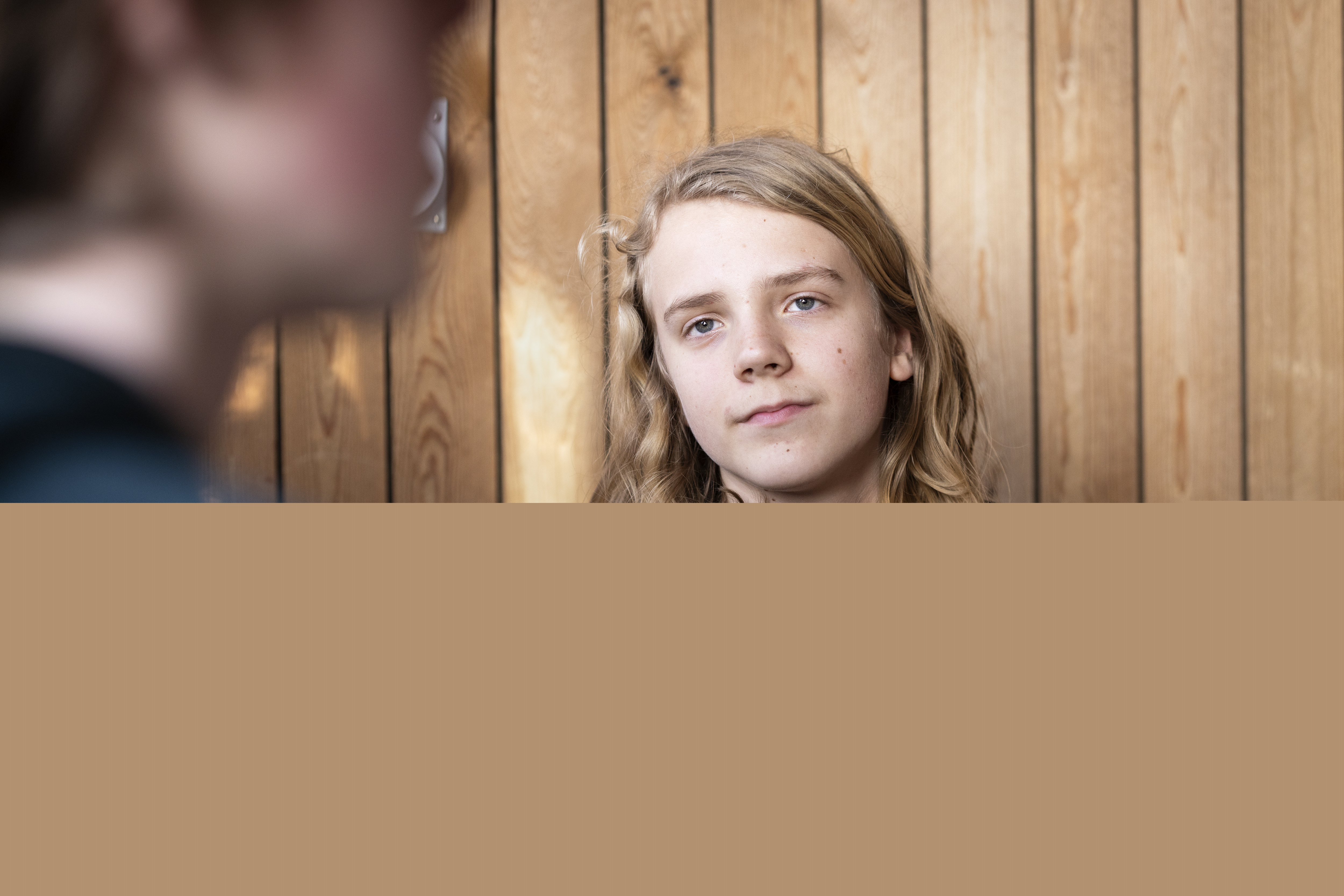 En ung kille med långt hår sitter i en soffa. Någon syns suddigt i förgrunden.