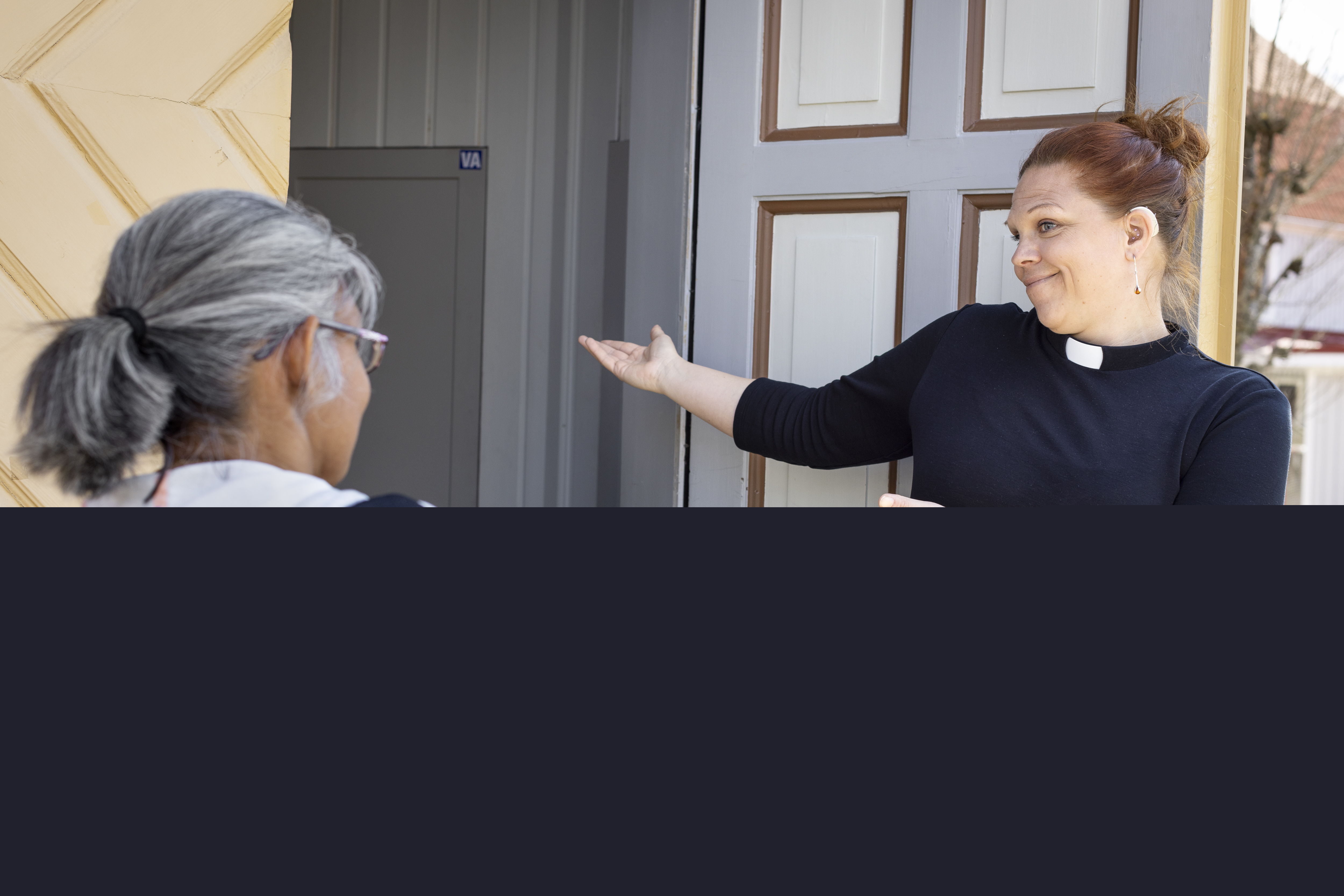 En kvinnlig präst visar vägen in i kyrkan med armarna åt en kvinna.