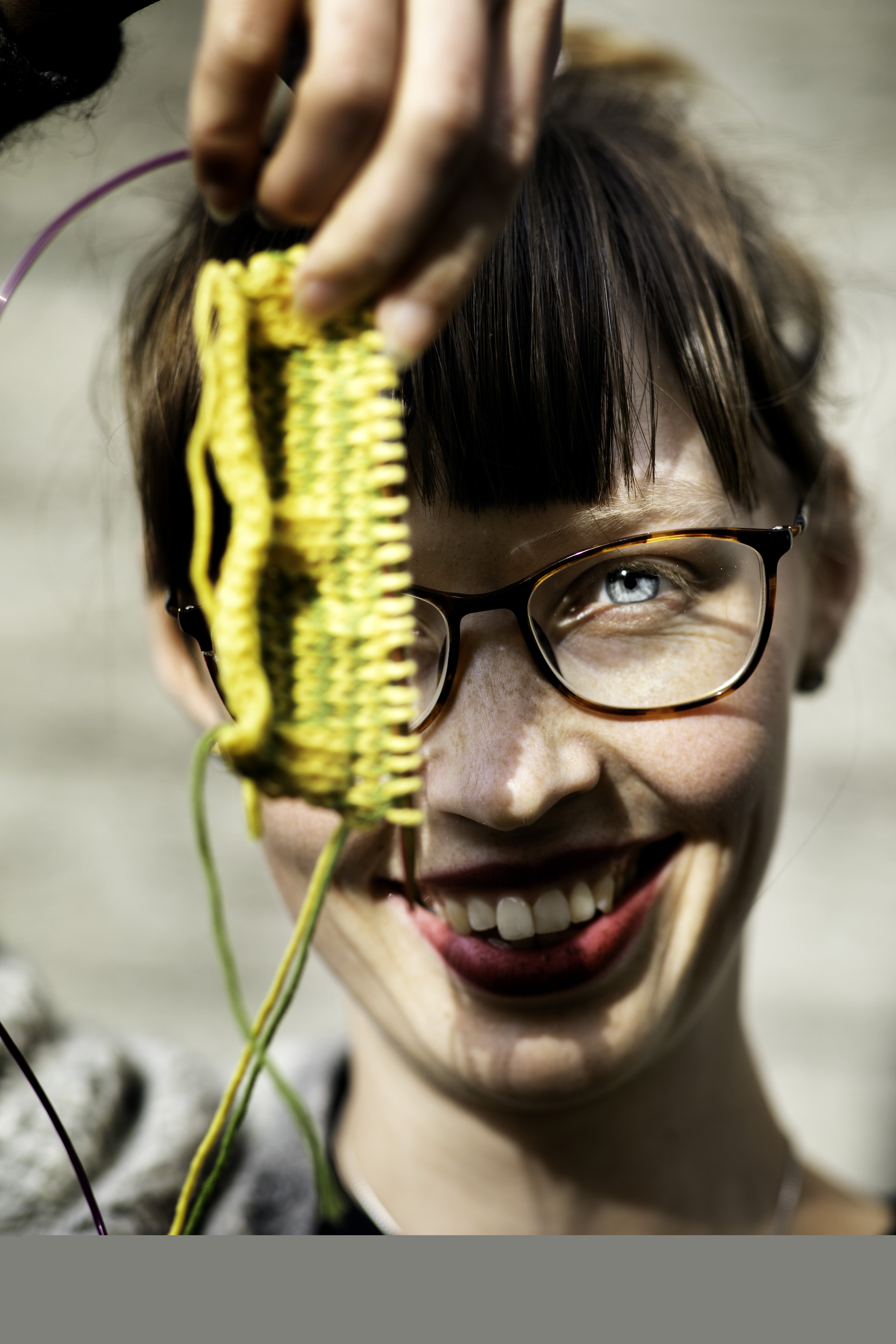 En kvinna med lugg och rött läppstift håller upp en gul stickning mot kameran och ler stort.