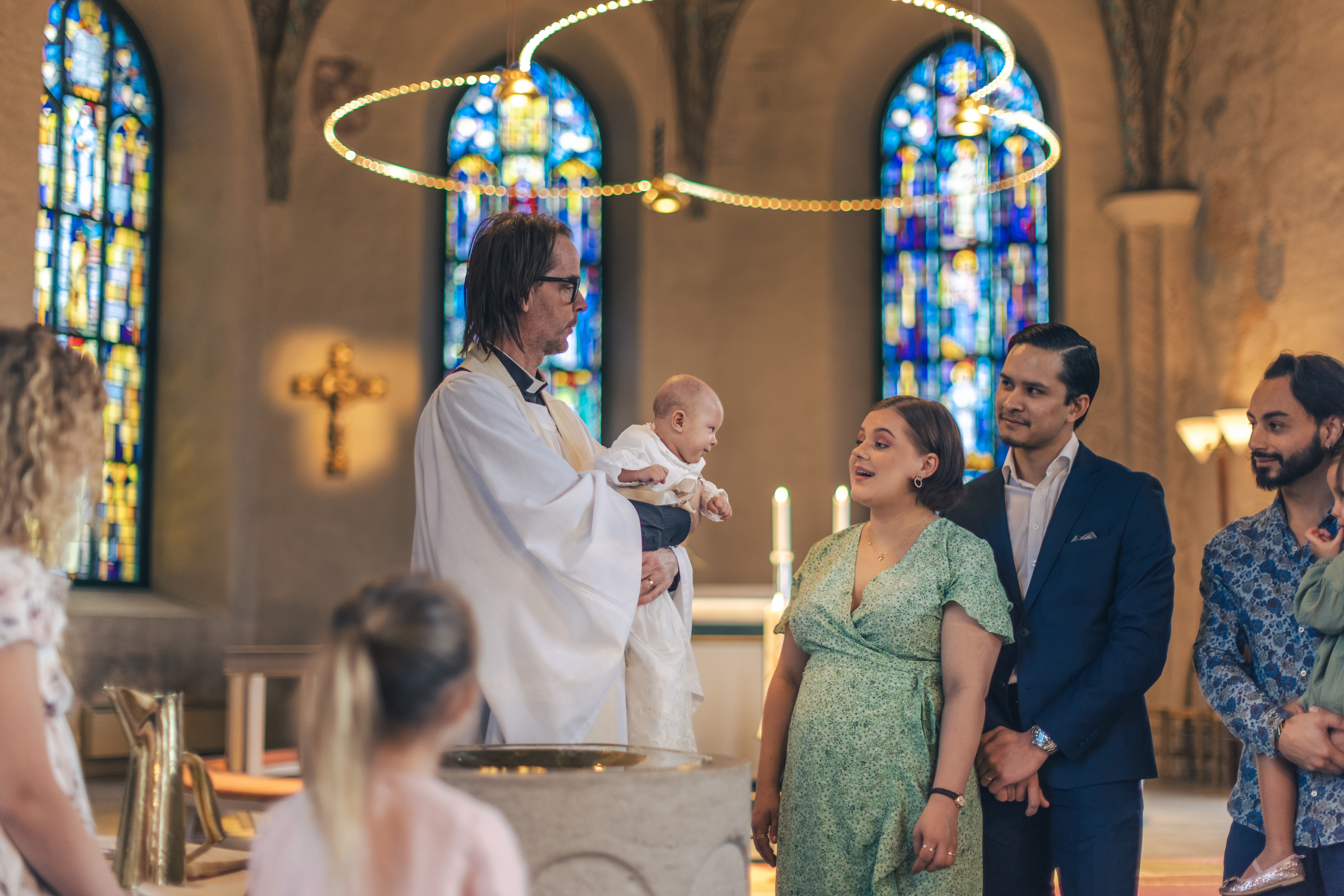 Ett dop i en kyrka. En kvinna och hennes man står bredvid prästen som håller barnet i famnen.