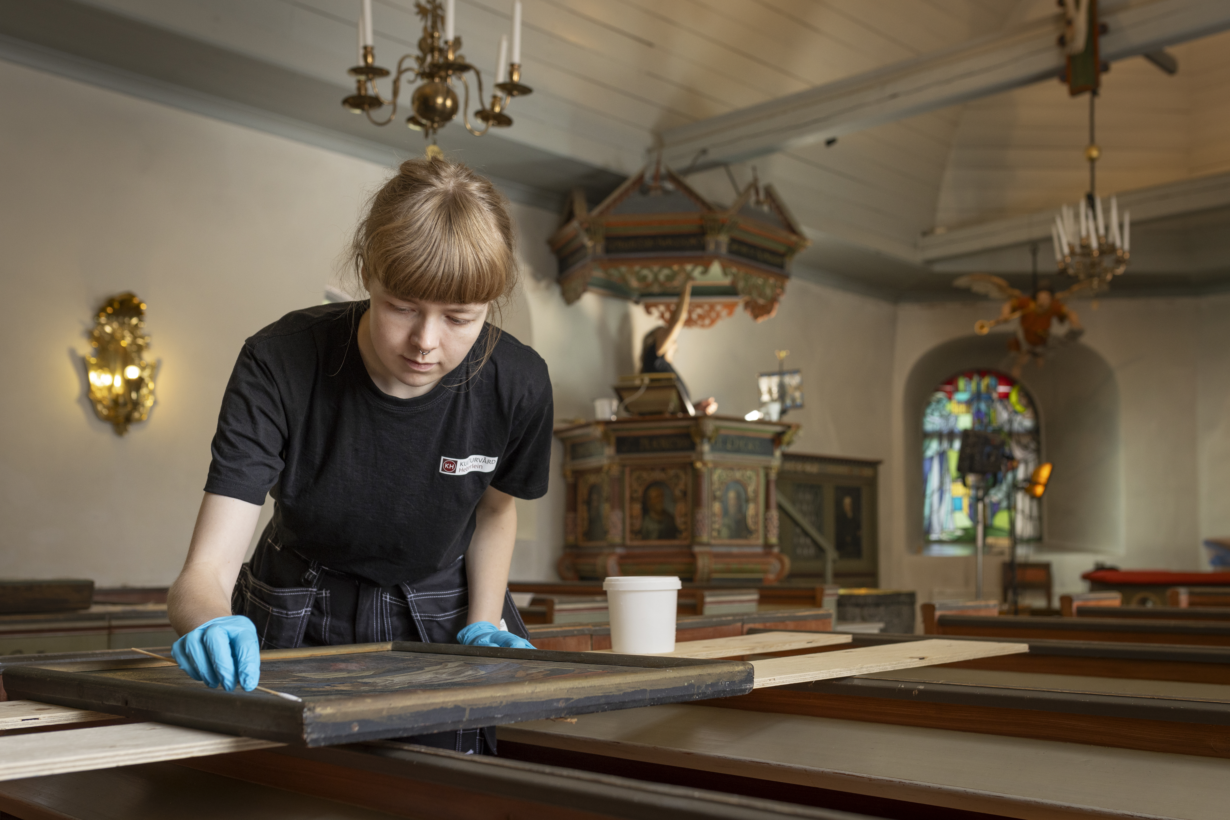 En kvinna i arbetskläder konserverar en tavla i an kyrka.
Fco