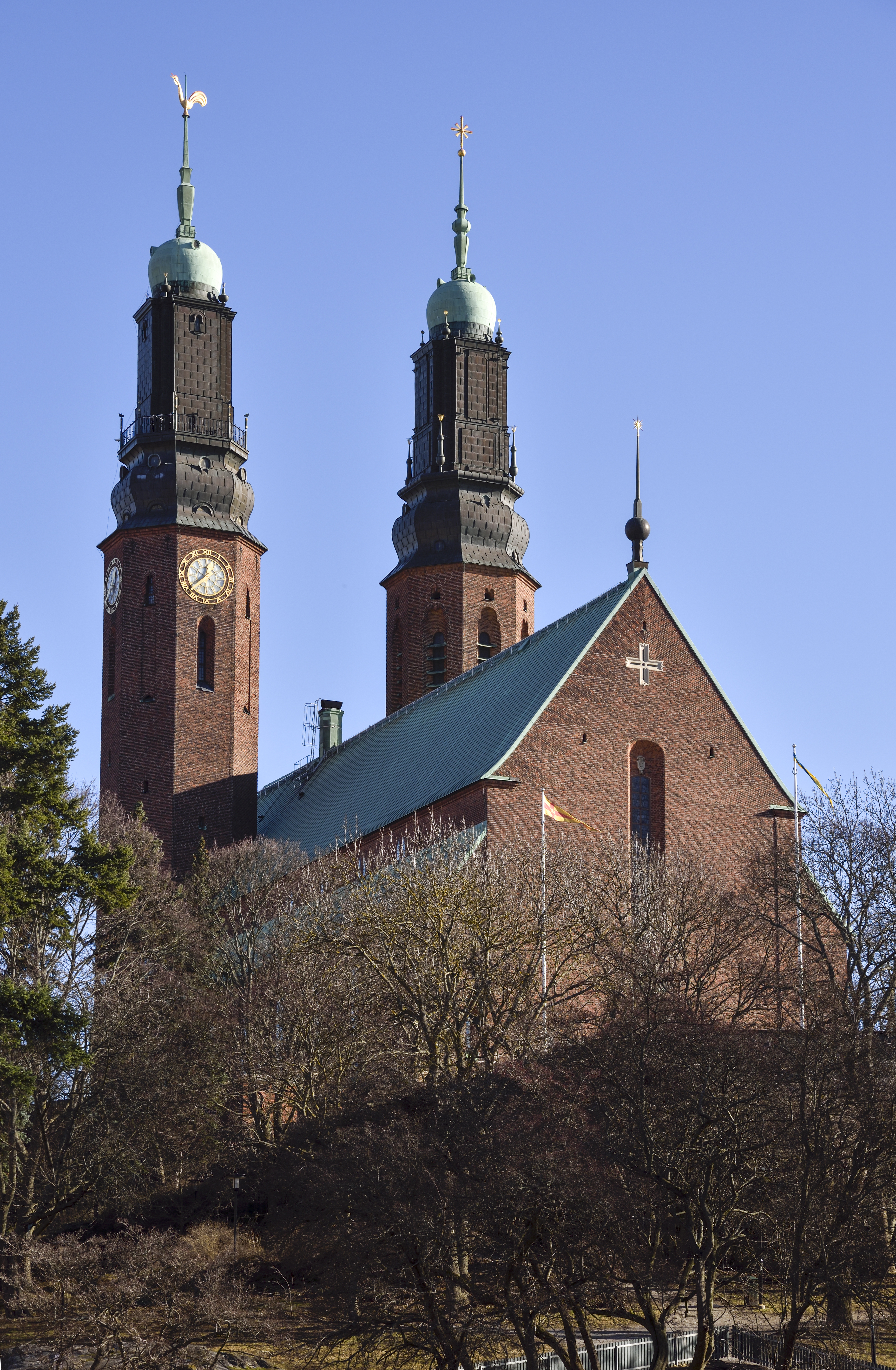 En röd tegelkyrka med dubbla torn. Klarblå himmel. Träd utan löv i förgrunden.