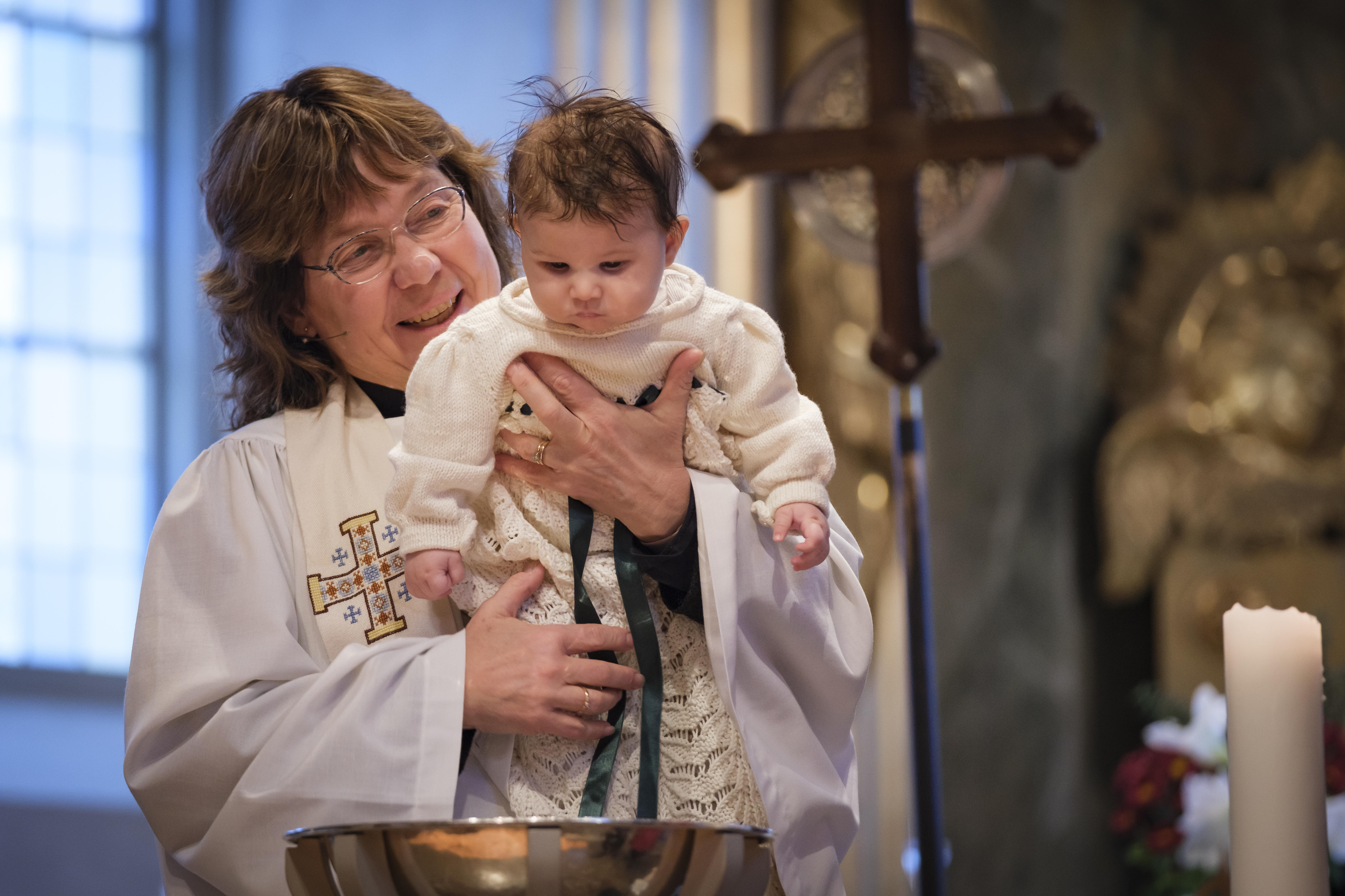 En kvinnlig präst håller en liten bebis i famnen ovanför dopfunten.