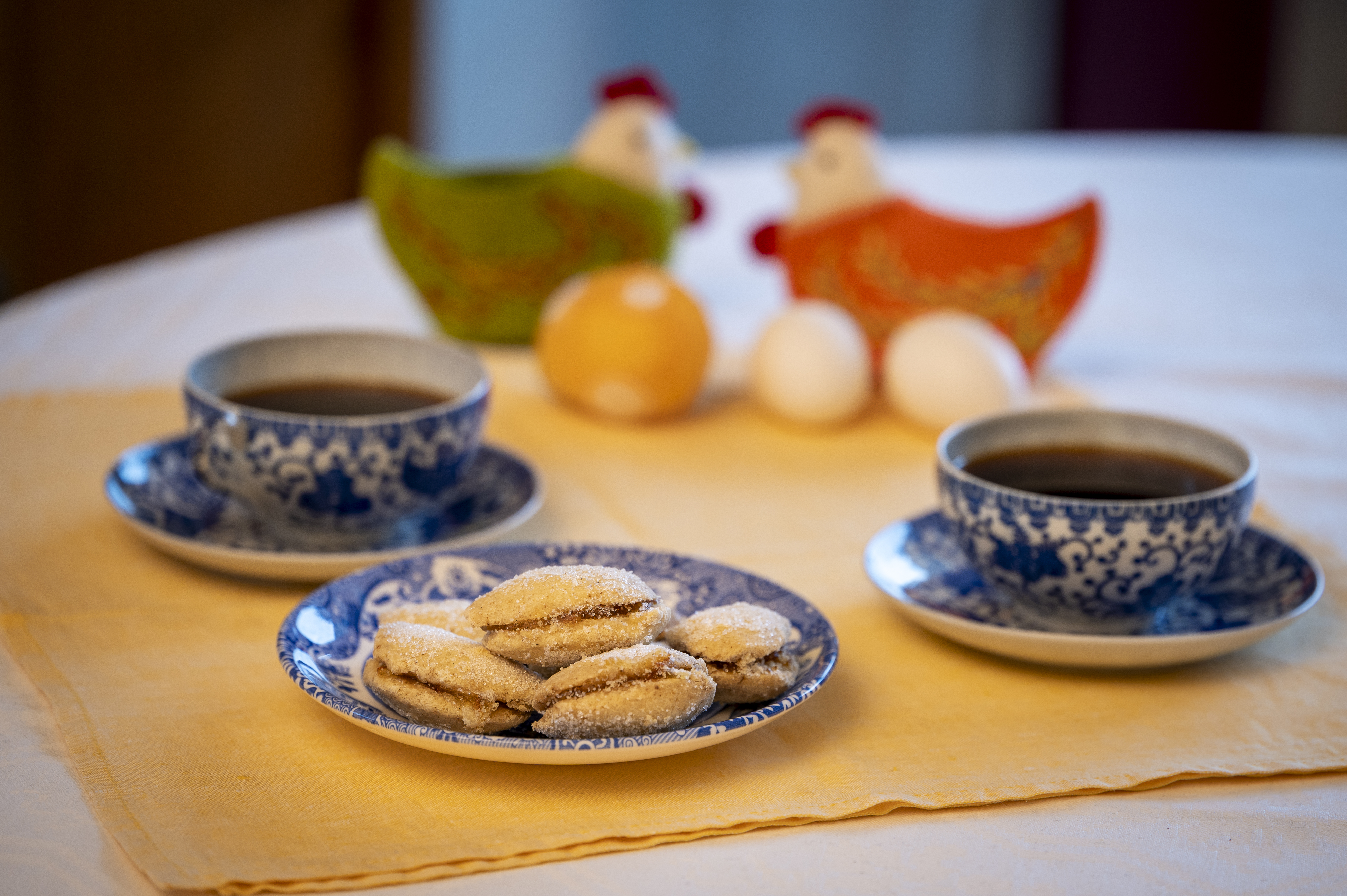 Ett kakfat, två kaffekoppar på fat och två påskhönor med ägg står uppdukade på en gul duk.