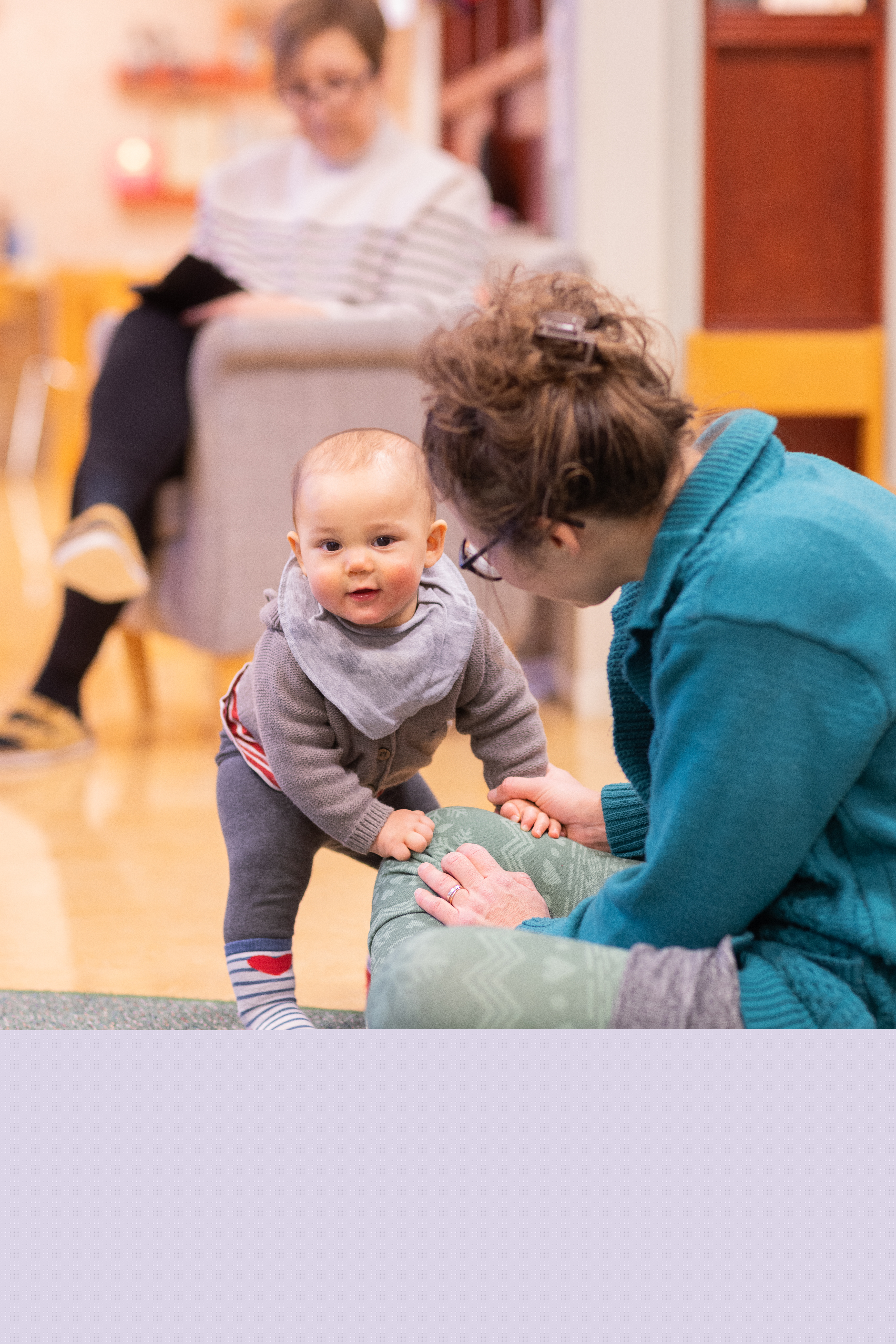 En bebis ställer sig trevande upp, stödd mot sin mamma.