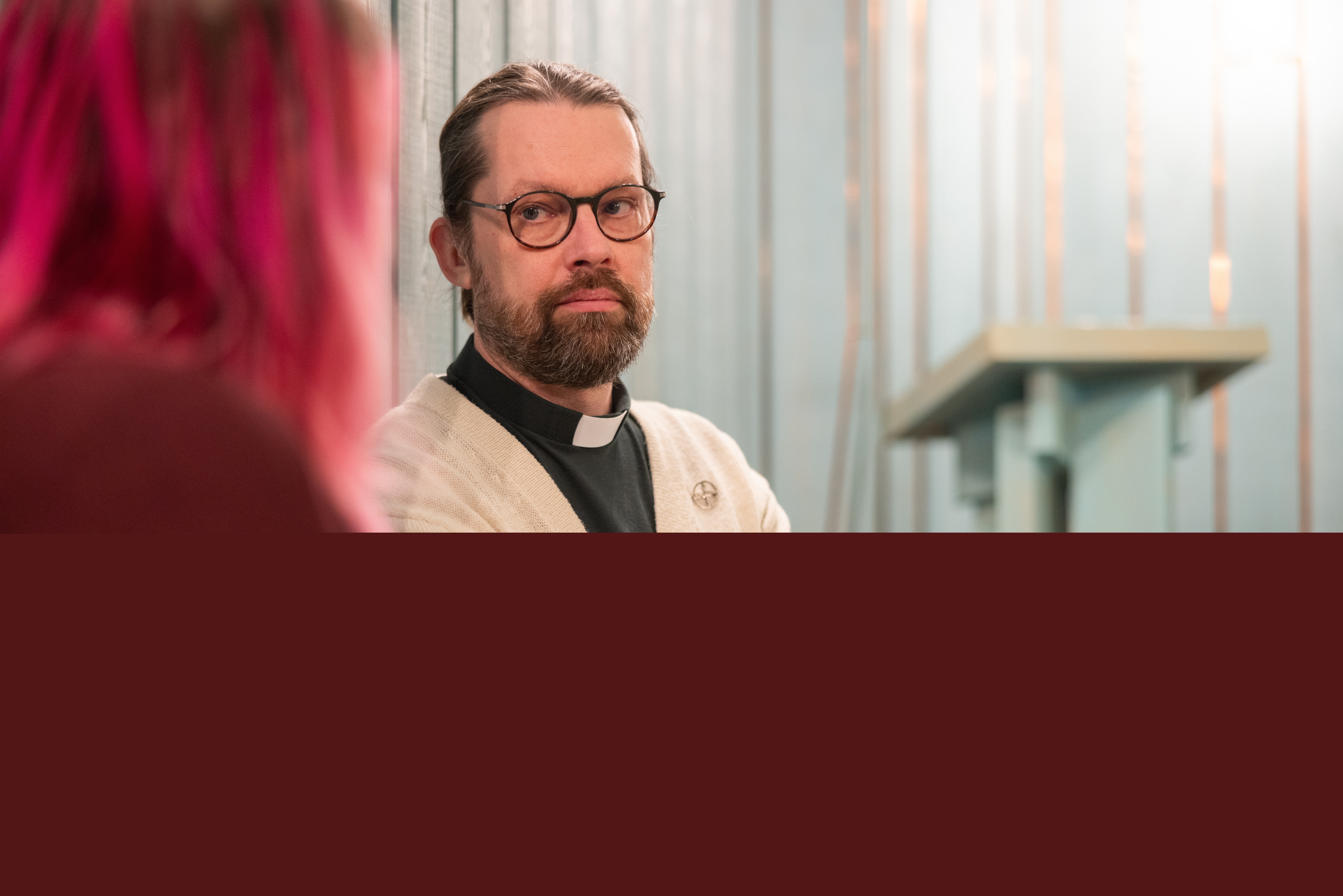 En diakon sitter inomhus och samtalar med en person med rosafärgat hår.