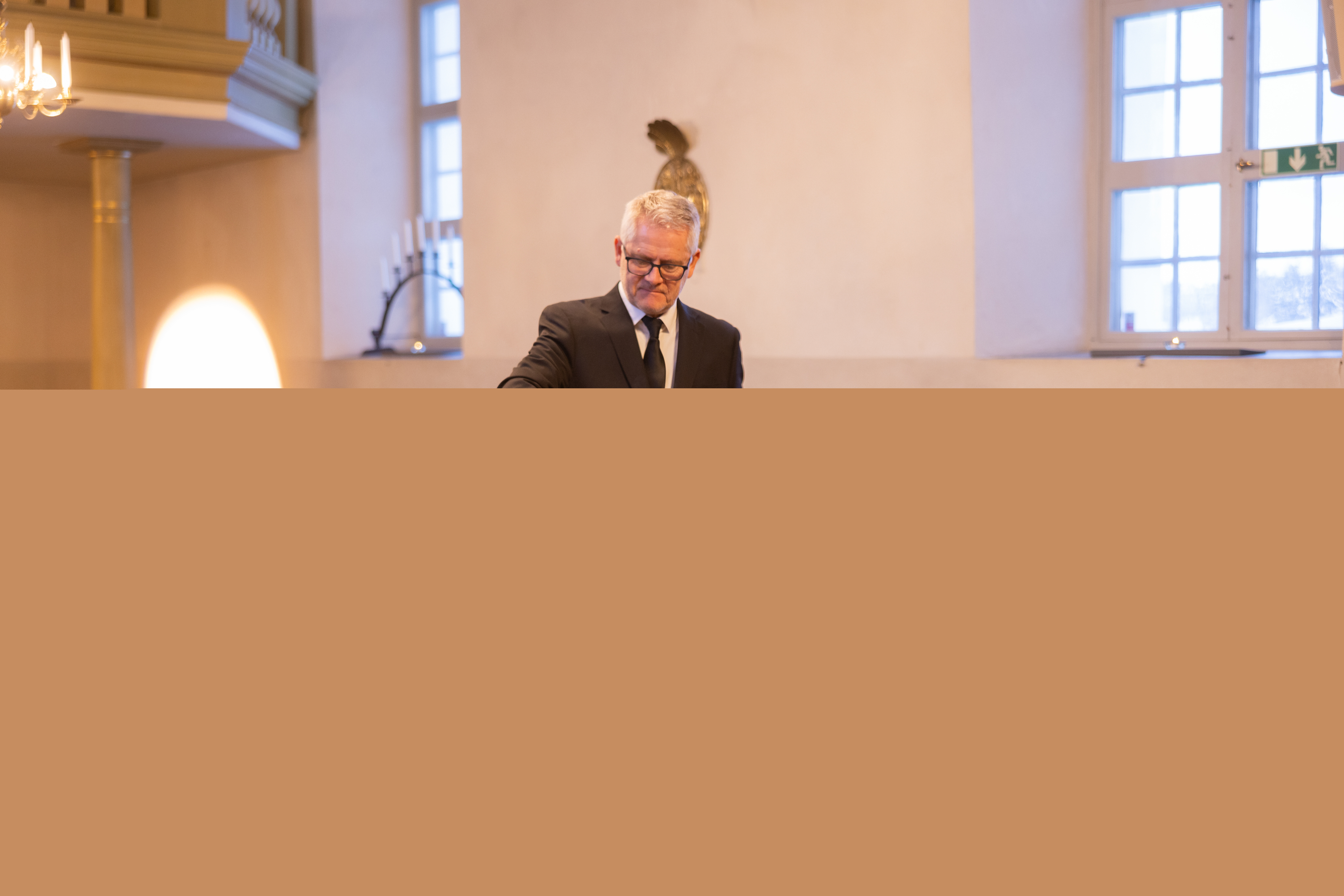 En kyrkvaktmästare tänder ett ljus på en vit kista som har en krans av amaryllis, tallkvistar och vita rosor.